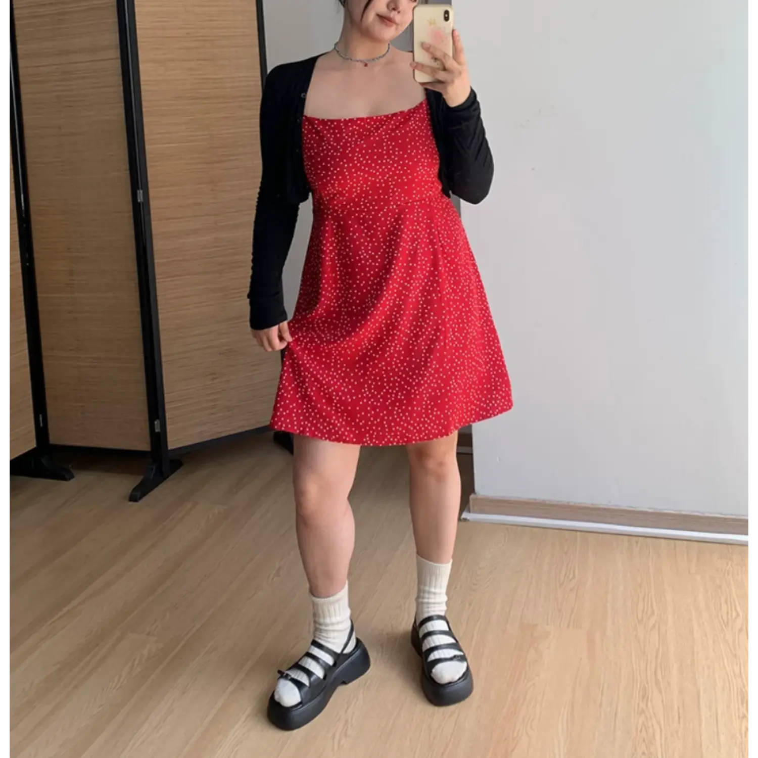 Medium full indoor mirror lifestyle shot of bright red micro polka dot mini dress, layered black fitted cropped knit cardigan, model resting one hand on hip to show waist silhouette
