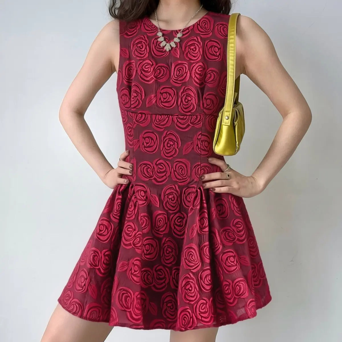 Woman styled in a red rose brocade sleeveless mini dress, featuring a fitted bodice and gathered flared skirt, accessorized with a yellow handbag.