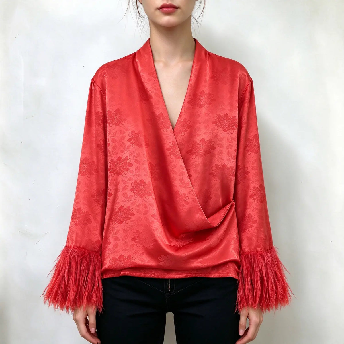 Model wearing red satin wrap blouse, subtle floral jacquard pattern, deep V wrap front, long sleeves with red feather fringe cuffs, relaxed fit, paired with black jeans, plain white background