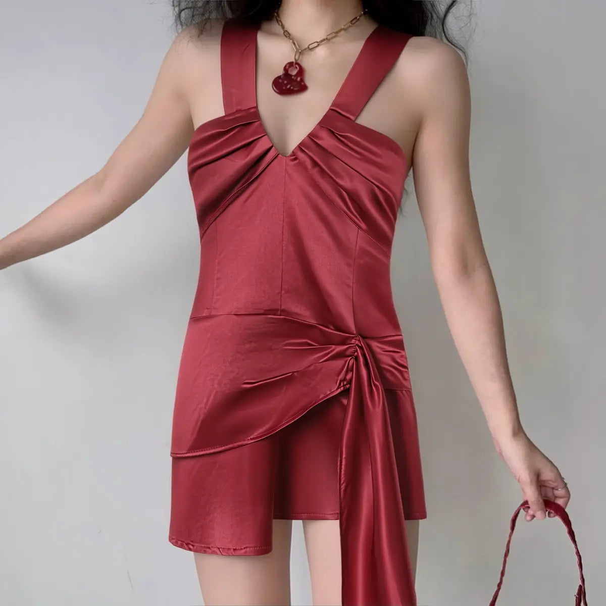 Model wearing a red satin mini dress with a gathered V-neck and draped side tie, styled with a yellow bag and butterfly necklace.