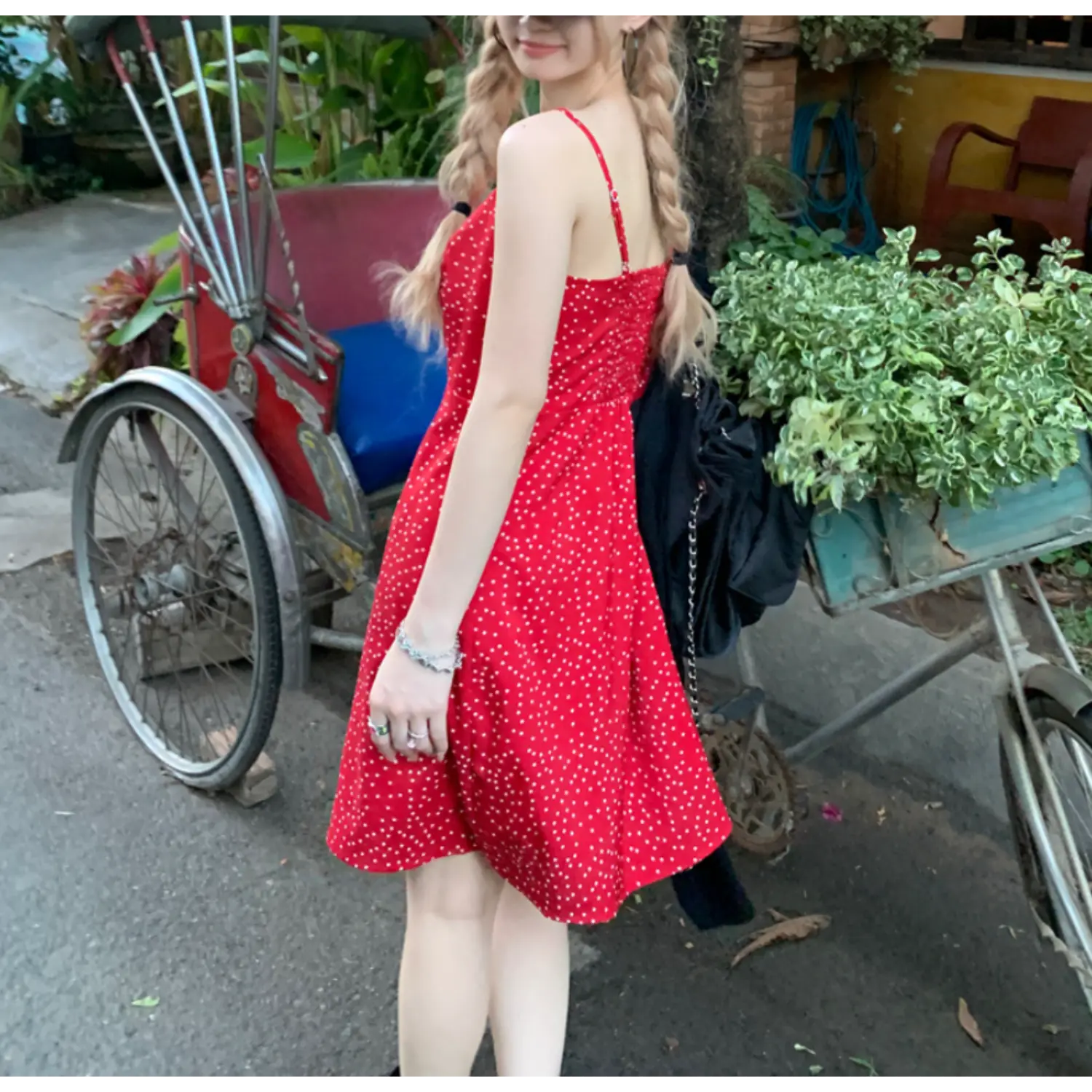 Outdoor travel shot of bright red polka dot mini slip dress, three-quarter back view beside vintage rickshaw and lush green foliage, black fabric crossbody bag slung over shoulder