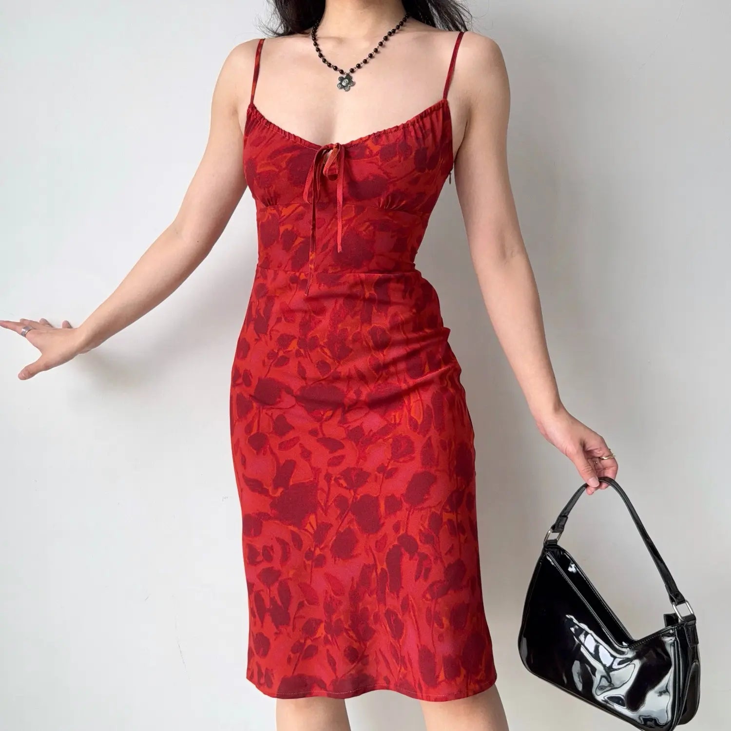 Lifestyle full-body shot of model wearing red abstract floral slip midi dress, thin adjustable spaghetti straps, bust self-tie drawstring, paired with black patent leather shoulder bag and beaded flower necklace