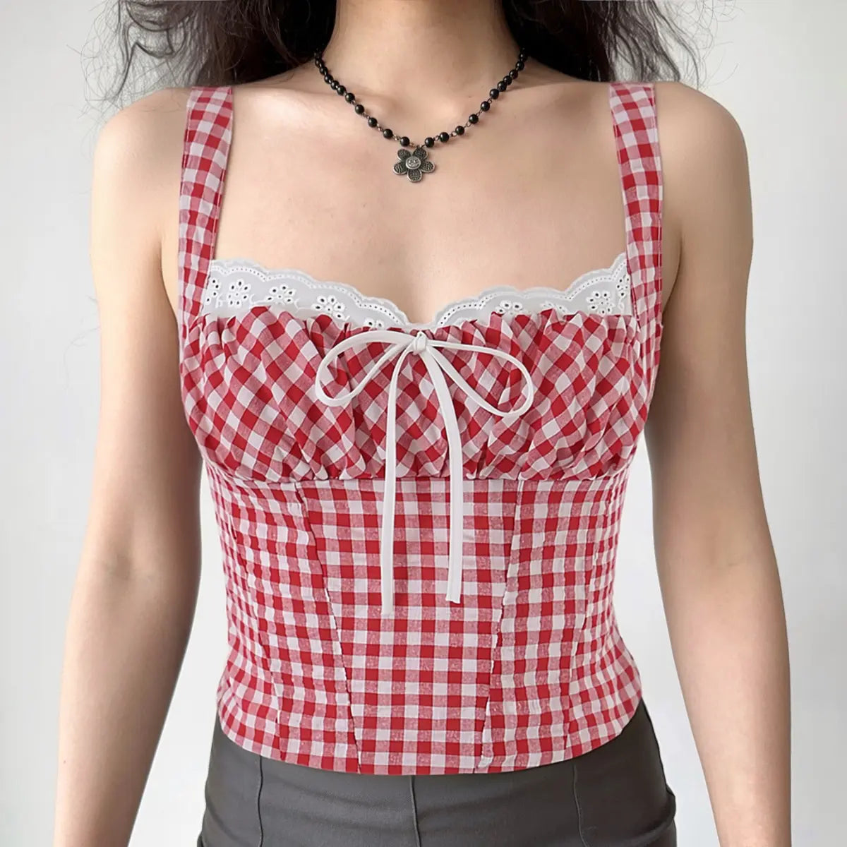 A woman wearing a red and white gingham bustier tank top with lace trim, center bow, and corset-style silhouette, styled with a black flower necklace and gray bottoms.