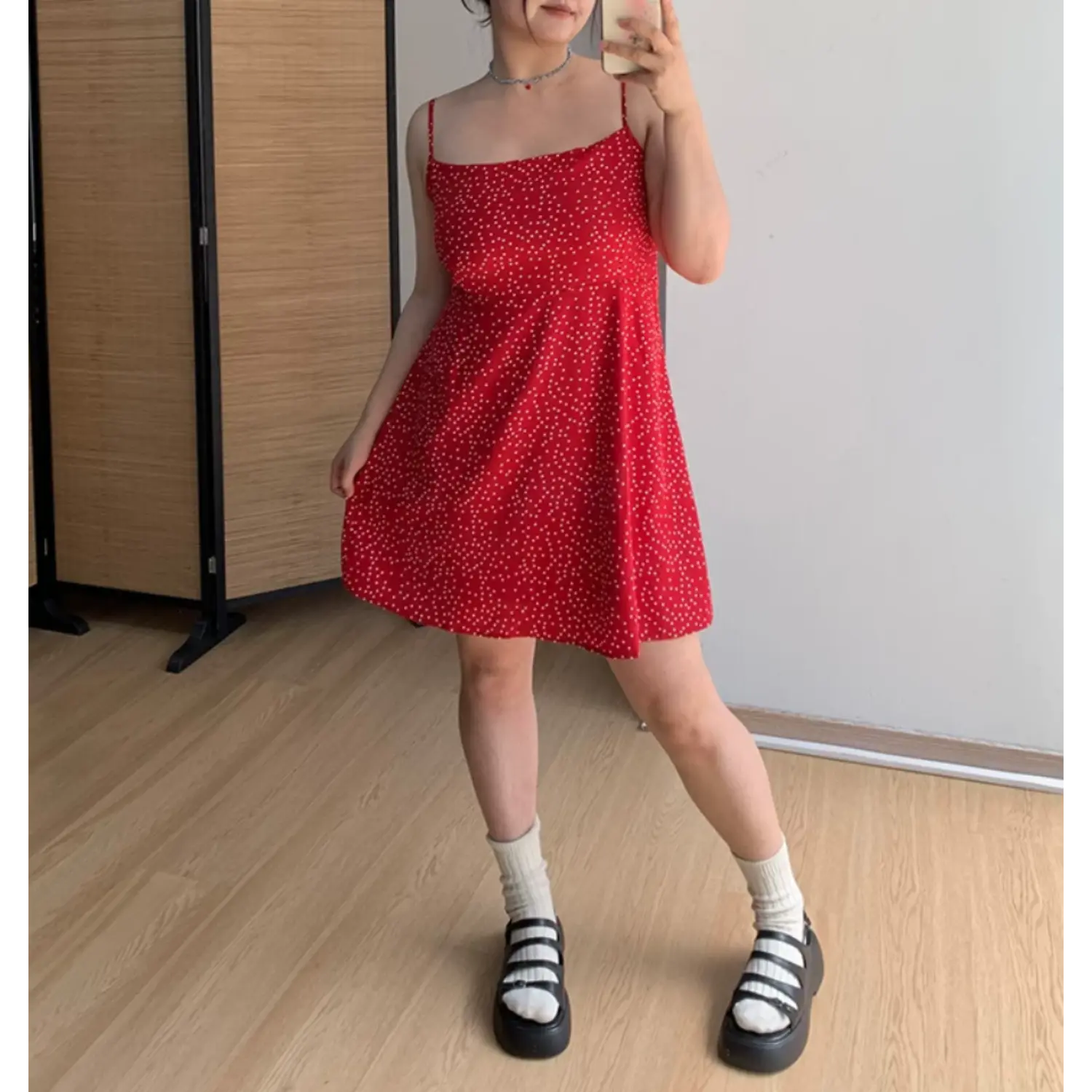 Angled indoor mirror lifestyle shot of red polka dot mini slip dress, model lifting skirt hem to show lightweight flowy fabric, white ribbed socks and black platform strappy shoes