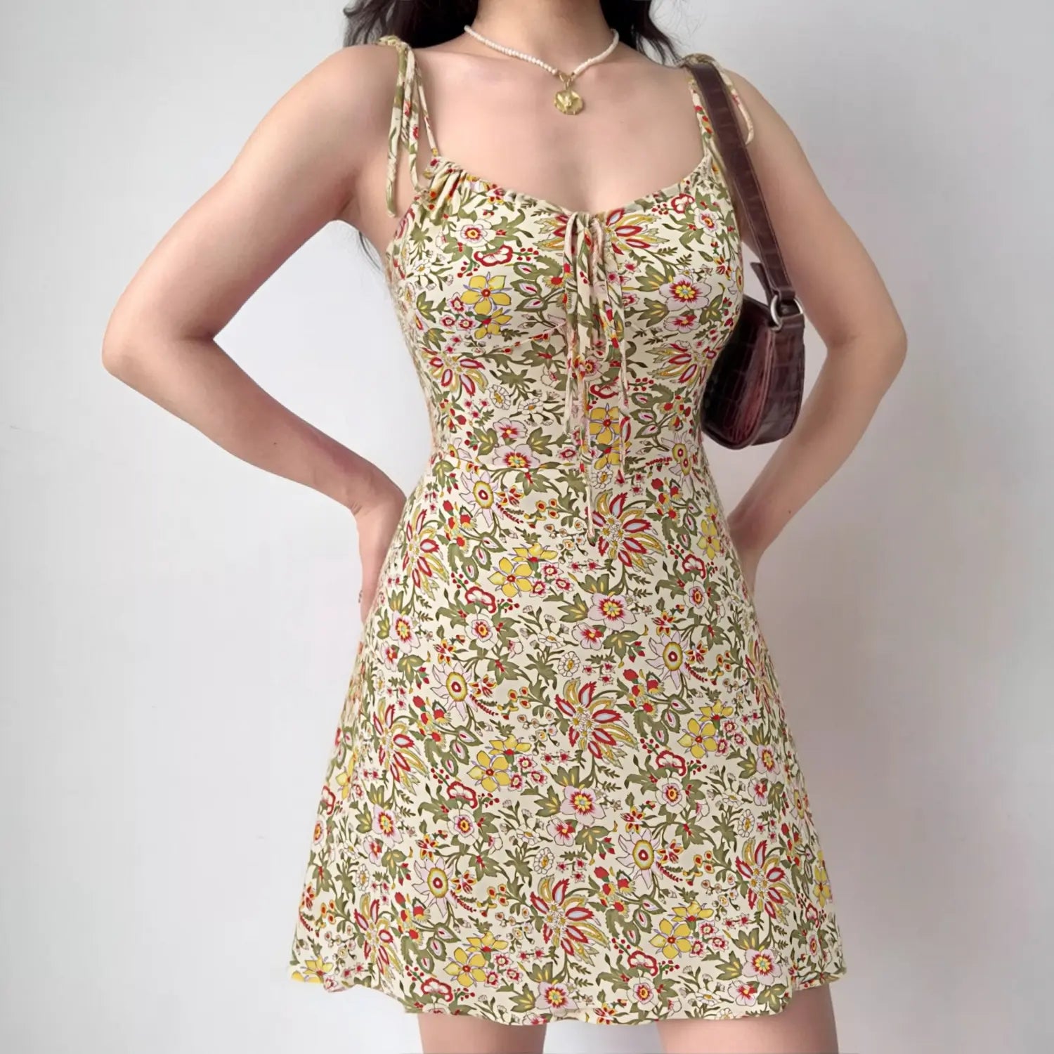 Indoor lifestyle shot of model wearing cream vintage floral tie strap mini slip dress, front bust drawstring bow, styled with brown saddle crossbody bag and dainty gold pendant necklace