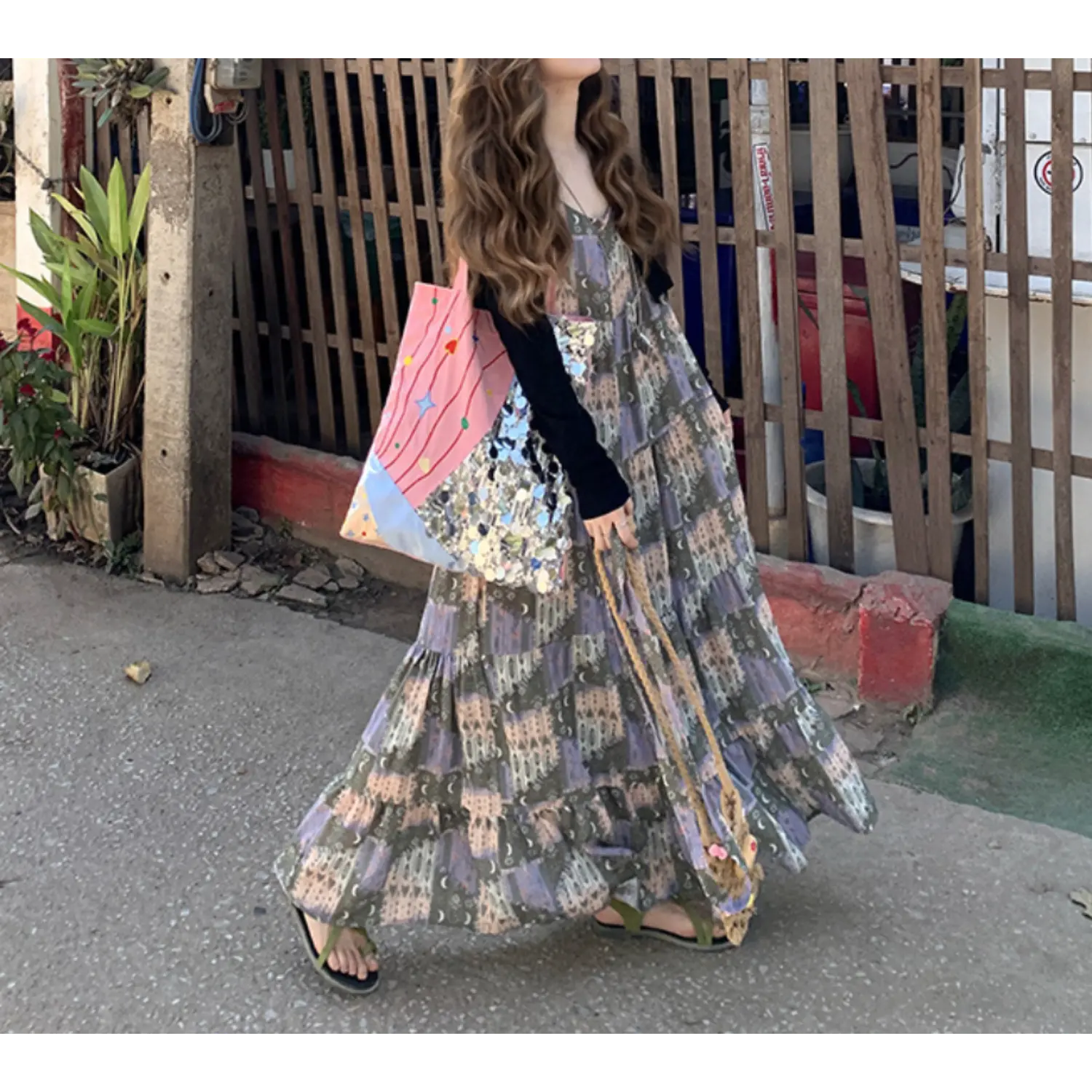 Three-quarter outdoor street lifestyle shot of whimsical printed tier slip maxi dress, vintage castle moon collage pattern, loose oversized tiered swing skirt, thin spaghetti straps, layered with long sleeve black knit cropped cardigan, green slide sandals, pink star graphic canvas tote bag, wooden slat fence & tropical roadside greenery backdrop