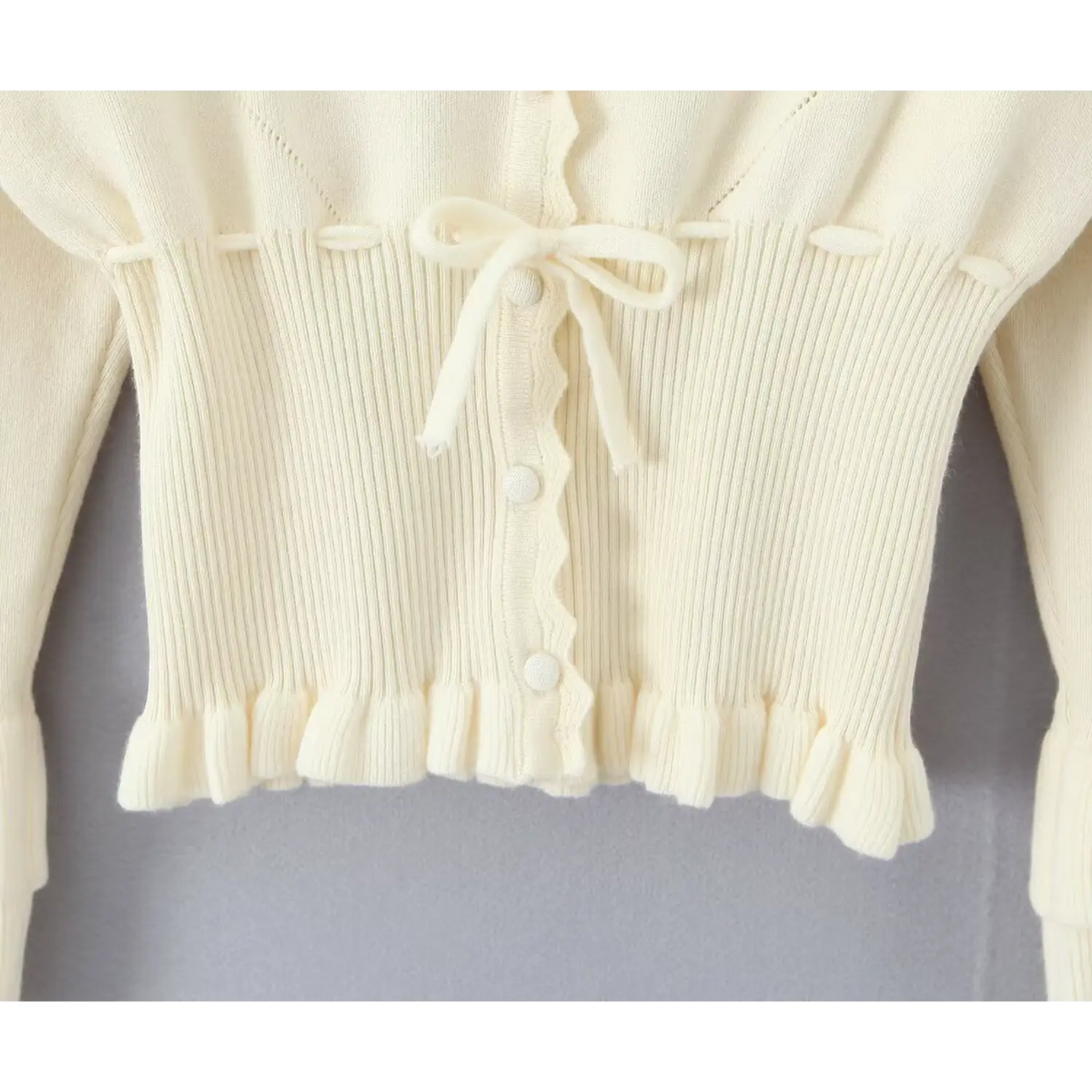 Close-up of scalloped placket with three round buttons, bow tie and ruffled hem on cream cardigan