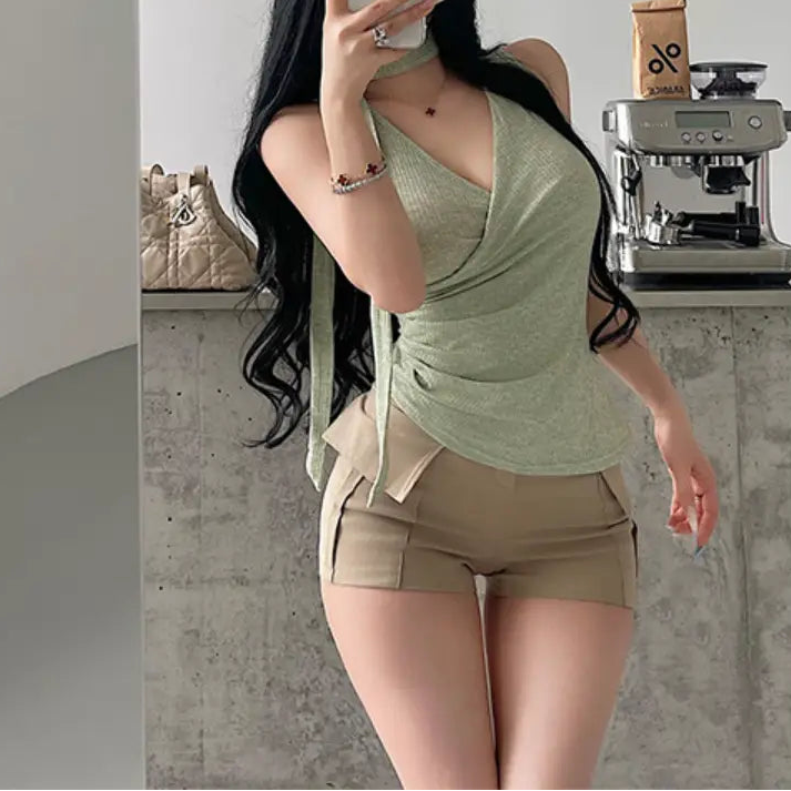 A woman wearing a sage green ribbed halter wrap top with a tie detail, styled with beige shorts, taking a mirror selfie.