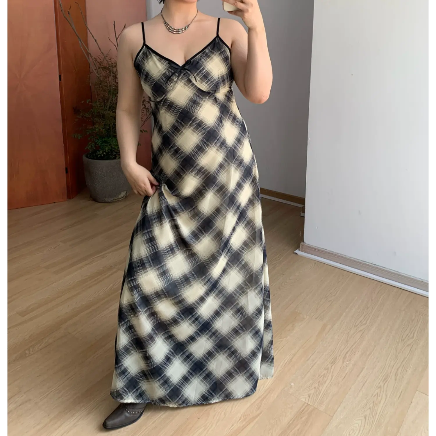 Front indoor mirror partial full shot of unlayered plaid slip maxi dress, contrast black piping along V-neck & straps, small chest fabric bow, bias cut diagonal check chiffon fabric, slim fit flared hem, minimalist light wood home interior