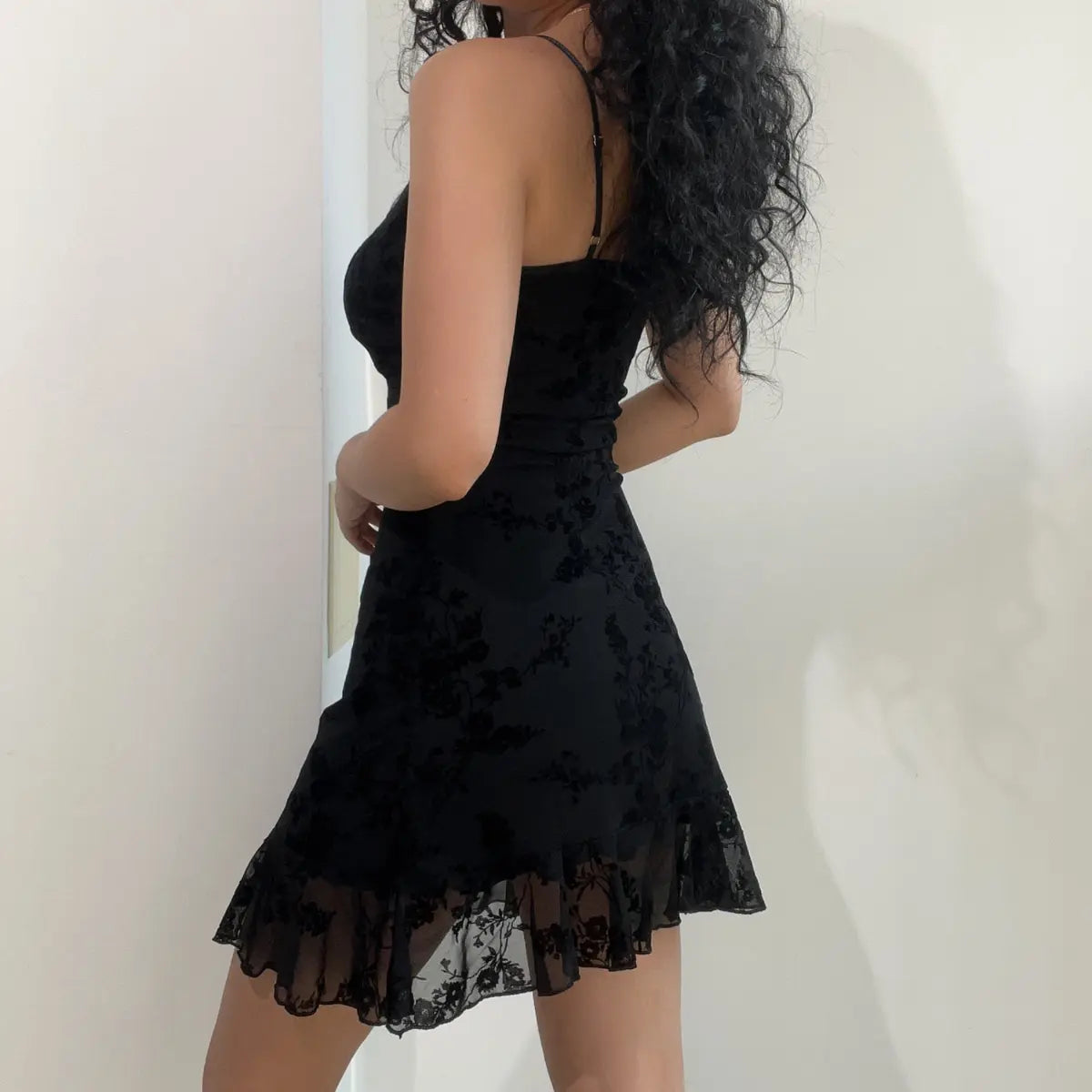 Side view of a black flocked floral mesh slip mini dress, showcasing the back strap adjuster and the flow of the sheer hem.