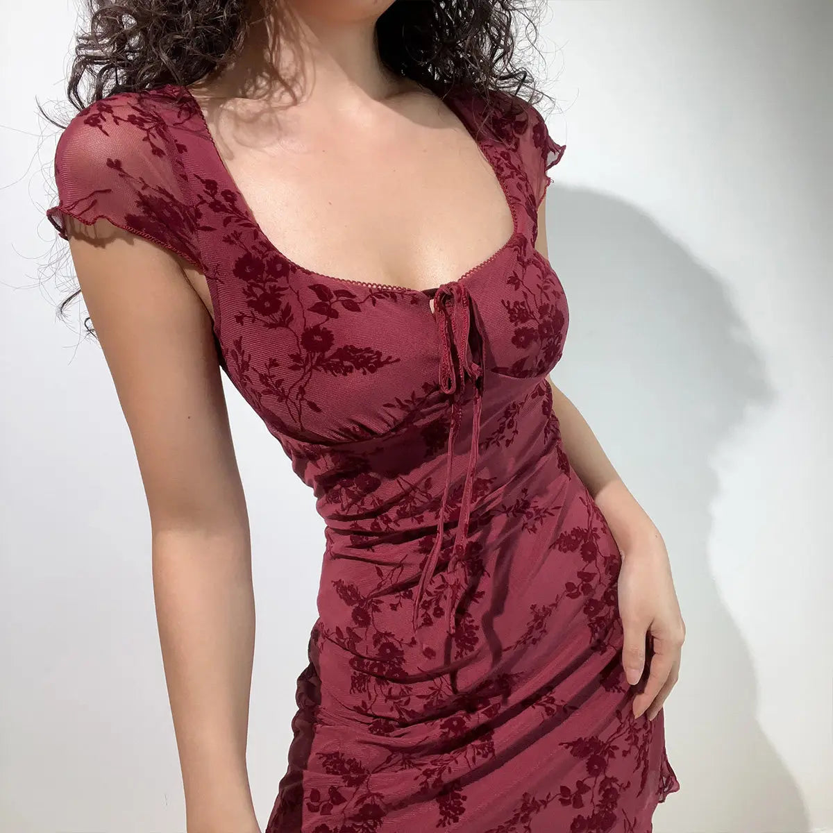  Side view of a burgundy flocked floral mesh mini dress, emphasizing the square neckline, cap sleeves, and bodycon fit.