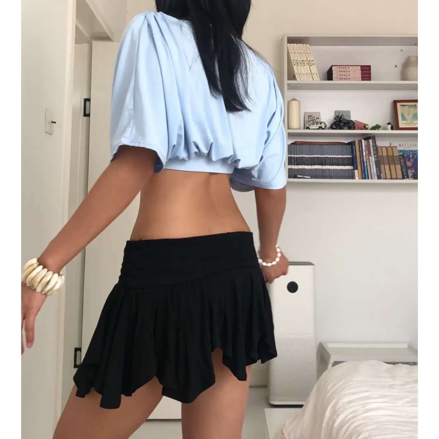 Back view indoor bedroom lifestyle shot of gathered surplice crop top, sky blue stretch jersey with full upper back ruching, loose batwing short sleeves, wide elastic cropped waist, styled with black low-rise ruffle mini skirt, stacked shell and pearl bracelets, white shelf background