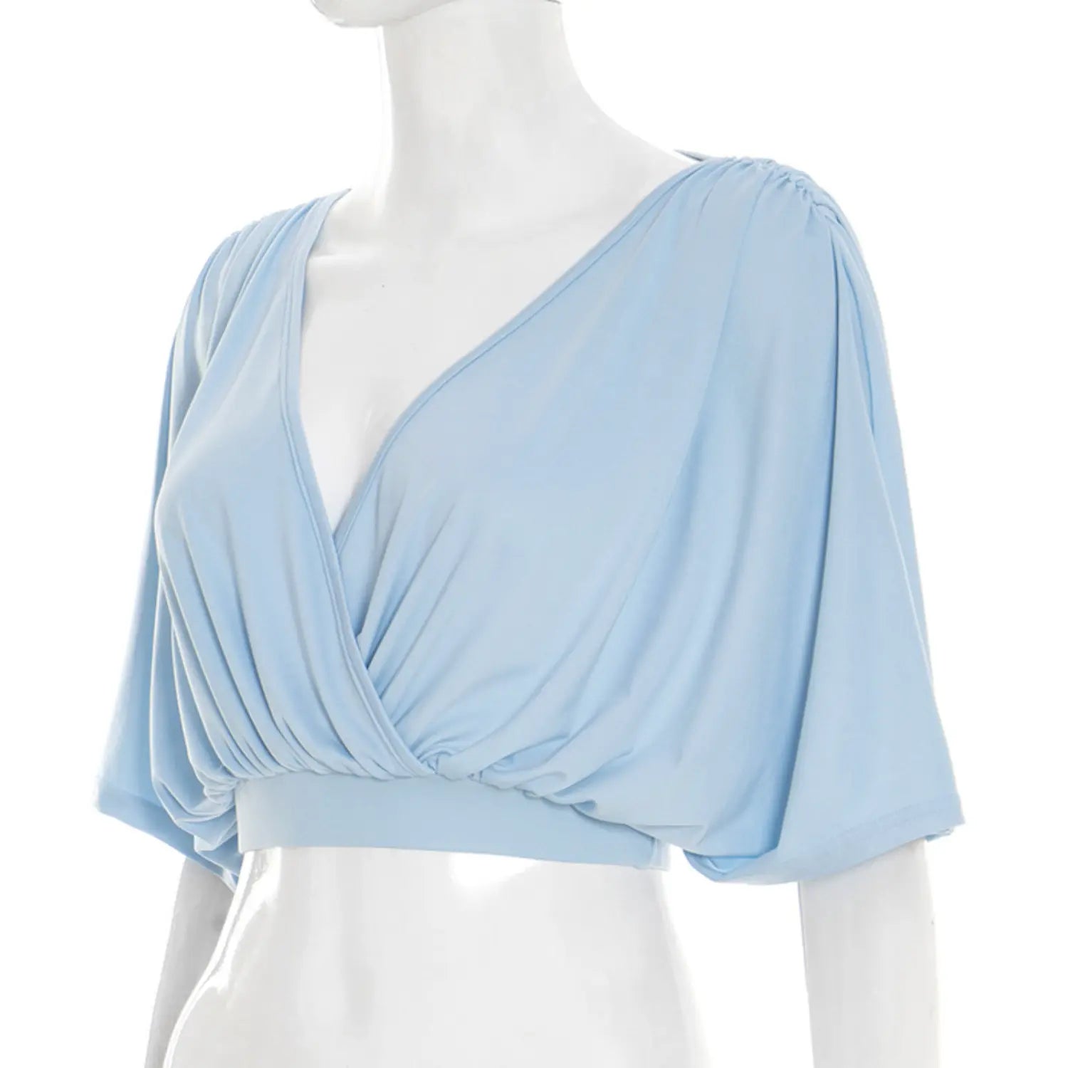 Three-quarter angled white studio mannequin shot of wrap cropped blouse, soft drapey sky blue jersey, crossover deep V neck, heavy front waist ruching, roomy batwing dolman sleeves, slim cropped elastic waist hem