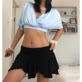 Minimalist indoor bedroom lifestyle shot of deep V wrap cropped blouse, soft sky blue stretch jersey with front shoulder ruching, oversized dolman short sleeves, styled with low-rise black ruffle mini skirt, layered silver star pendant necklaces, chunky shell bangle bracelets against white bookshelf backdrop