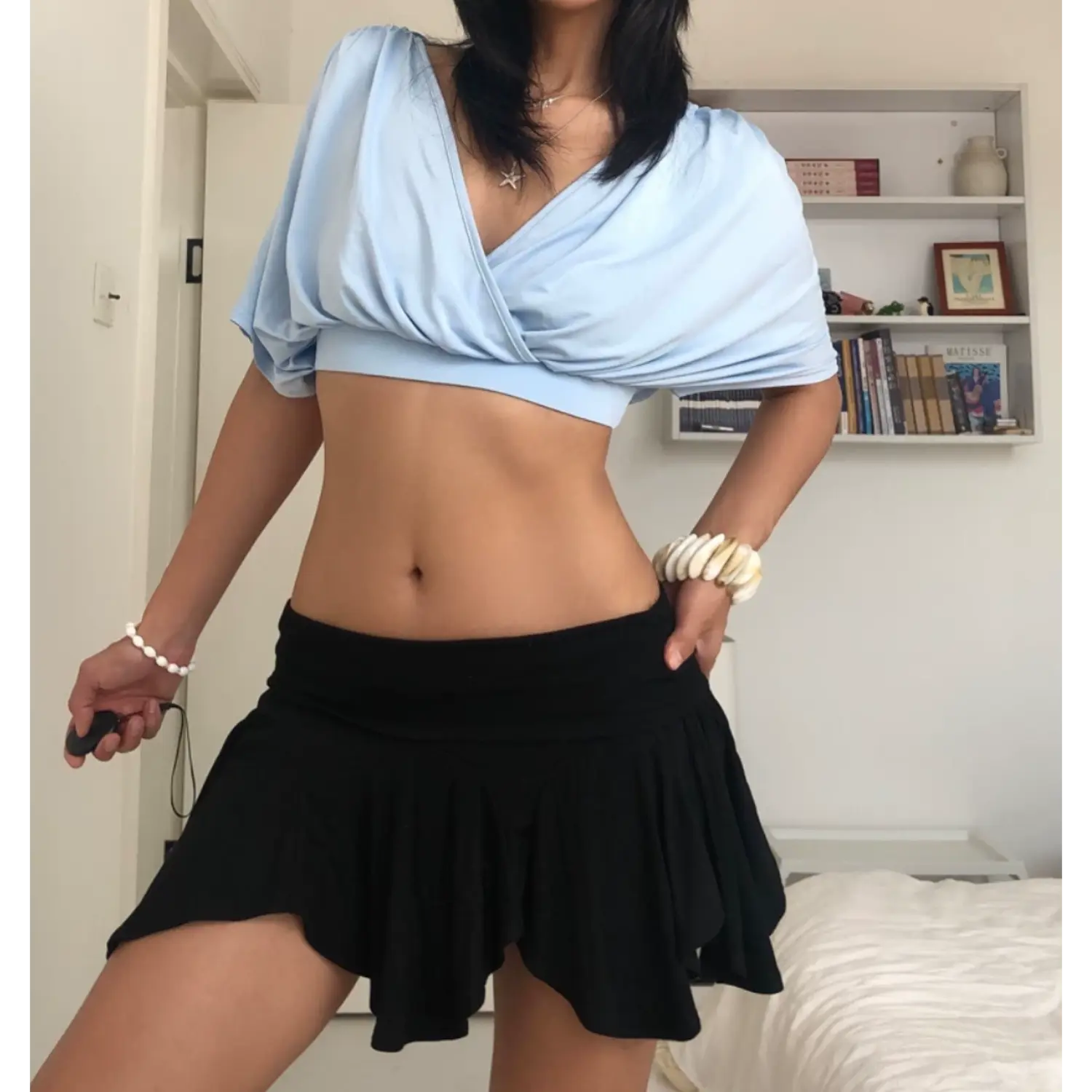 Minimalist indoor bedroom lifestyle shot of deep V wrap cropped blouse, soft sky blue stretch jersey with front shoulder ruching, oversized dolman short sleeves, styled with low-rise black ruffle mini skirt, layered silver star pendant necklaces, chunky shell bangle bracelets against white bookshelf backdrop