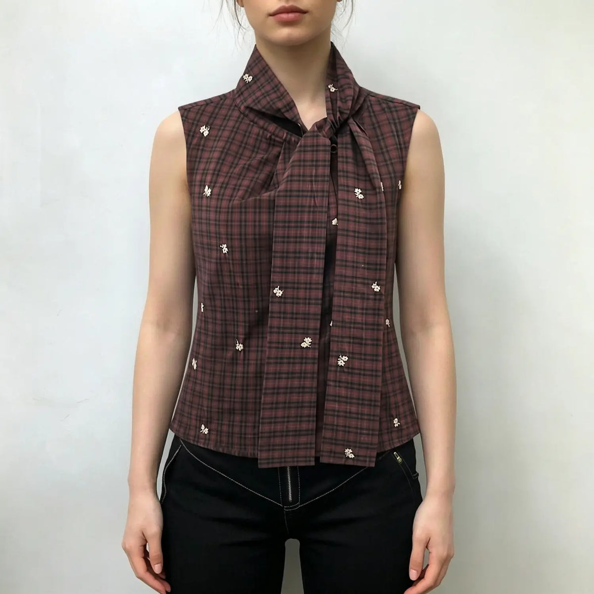Model wearing sleeveless plaid vest with tie front and floral embroidery, paired with black slim trousers on white background