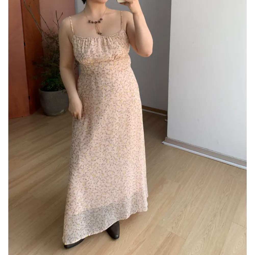 Indoor mirror full shot of pale peach micro floral spaghetti strap maxi dress, smocked elastic bust waist, flowy sheer floor-length skirt, paired with dark brown pointed leather ankle boots