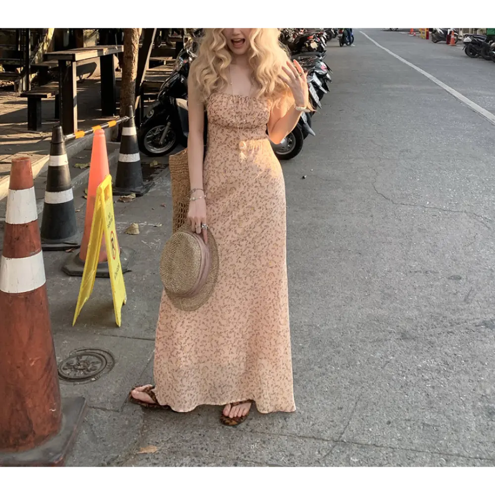 Full body urban street lifestyle shot of pale apricot micro floral spaghetti strap maxi dress, model waving one hand, holding woven straw sun hat, paired with leopard slide sandals and rattan crossbody bag