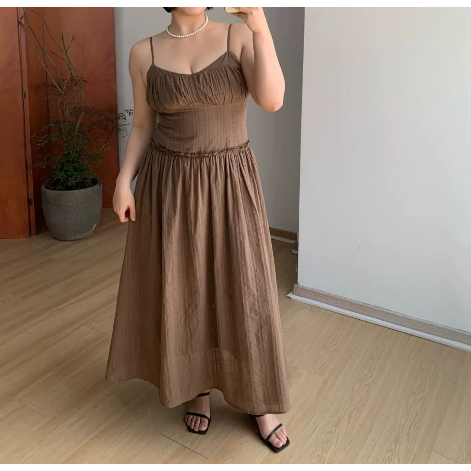 Casual front indoor mirror full shot of mocha brown textured cami maxi dress, ruched sweetheart bust, ruffled empire waist, voluminous gathered flowy long skirt, thin adjustable spaghetti straps, lightweight soft matte fabric, black thin strap slide sandals, minimalist neutral room decor