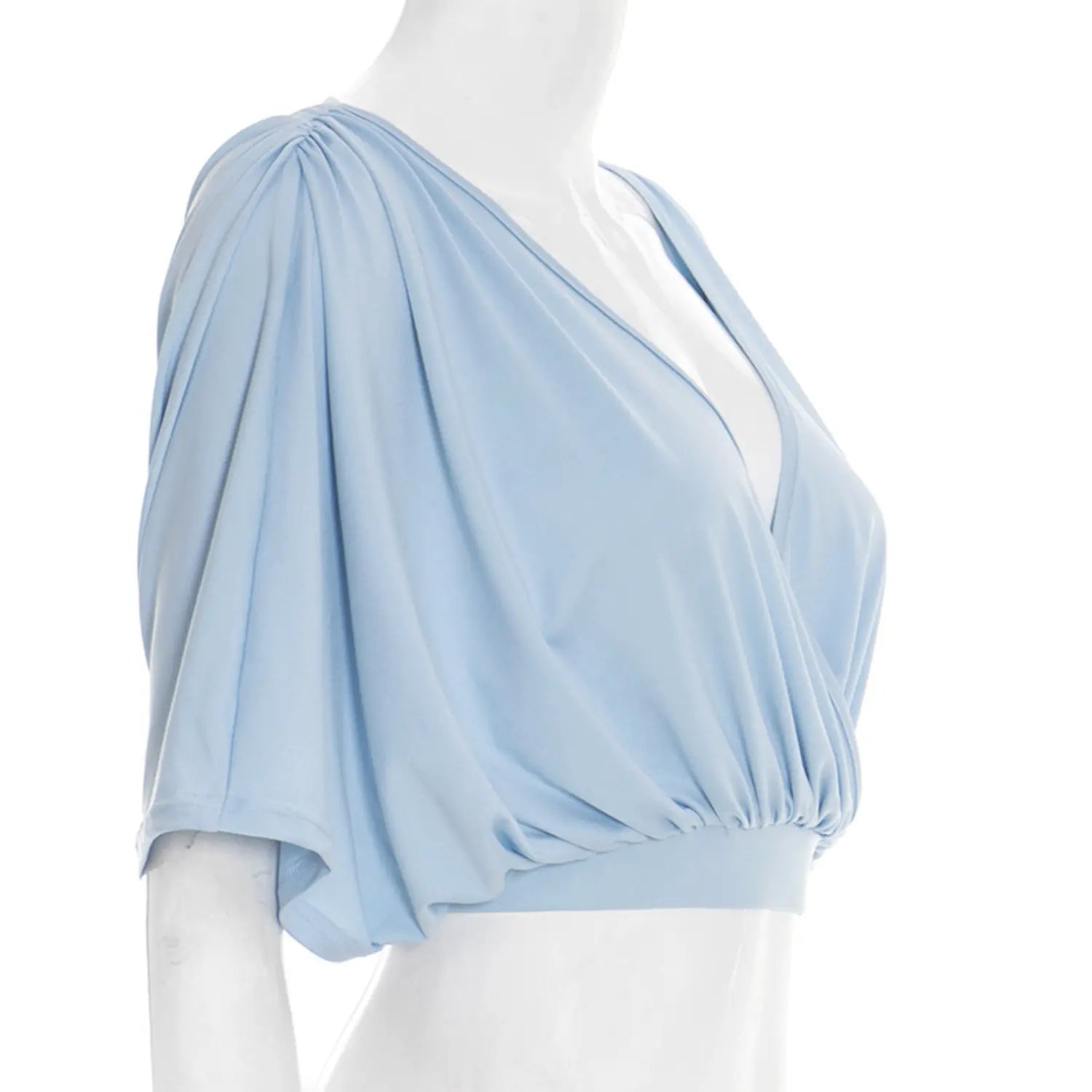 Side profile white studio mannequin shot of surplice wrap cropped blouse, deep V crossover neck, voluminous loose batwing dolman short sleeves, full front waist ruching, wide sky blue elastic cropped waistband
