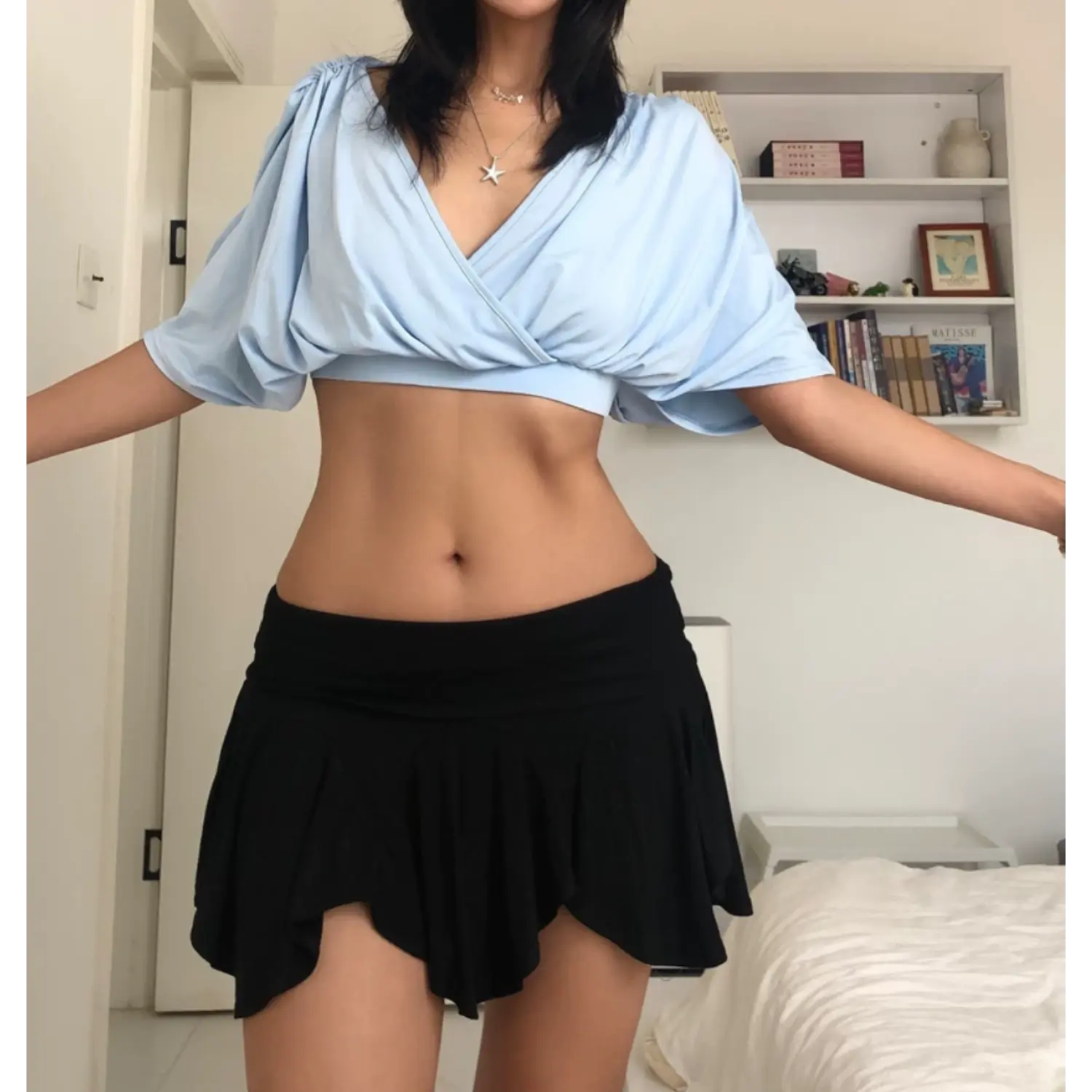 Relaxed full indoor bedroom shot of sky blue crossover V-neck cropped top, model stretching both arms out wide to display voluminous dolman sleeves, paired with black asymmetric ruffle mini skirt, dainty layered silver necklaces, bright minimalist white home interior