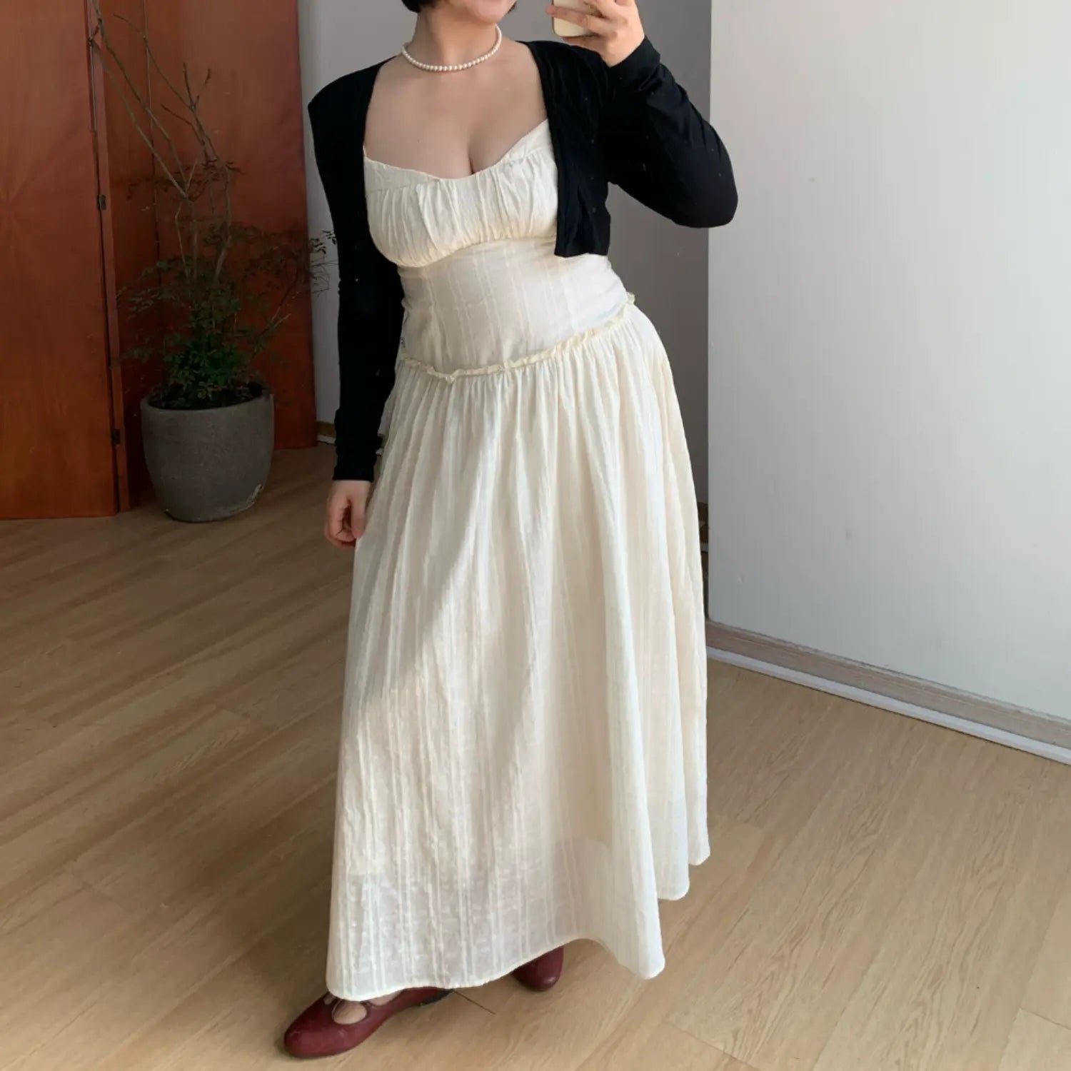 Three-quarter indoor mirror lifestyle shot of textured empire waist maxi slip dress layered with black cropped cardigan, model standing relaxed to show waist shaping & full skirt drape, subtle vertical waffle fabric, burgundy flat shoes, plain white wall & potted plant decor