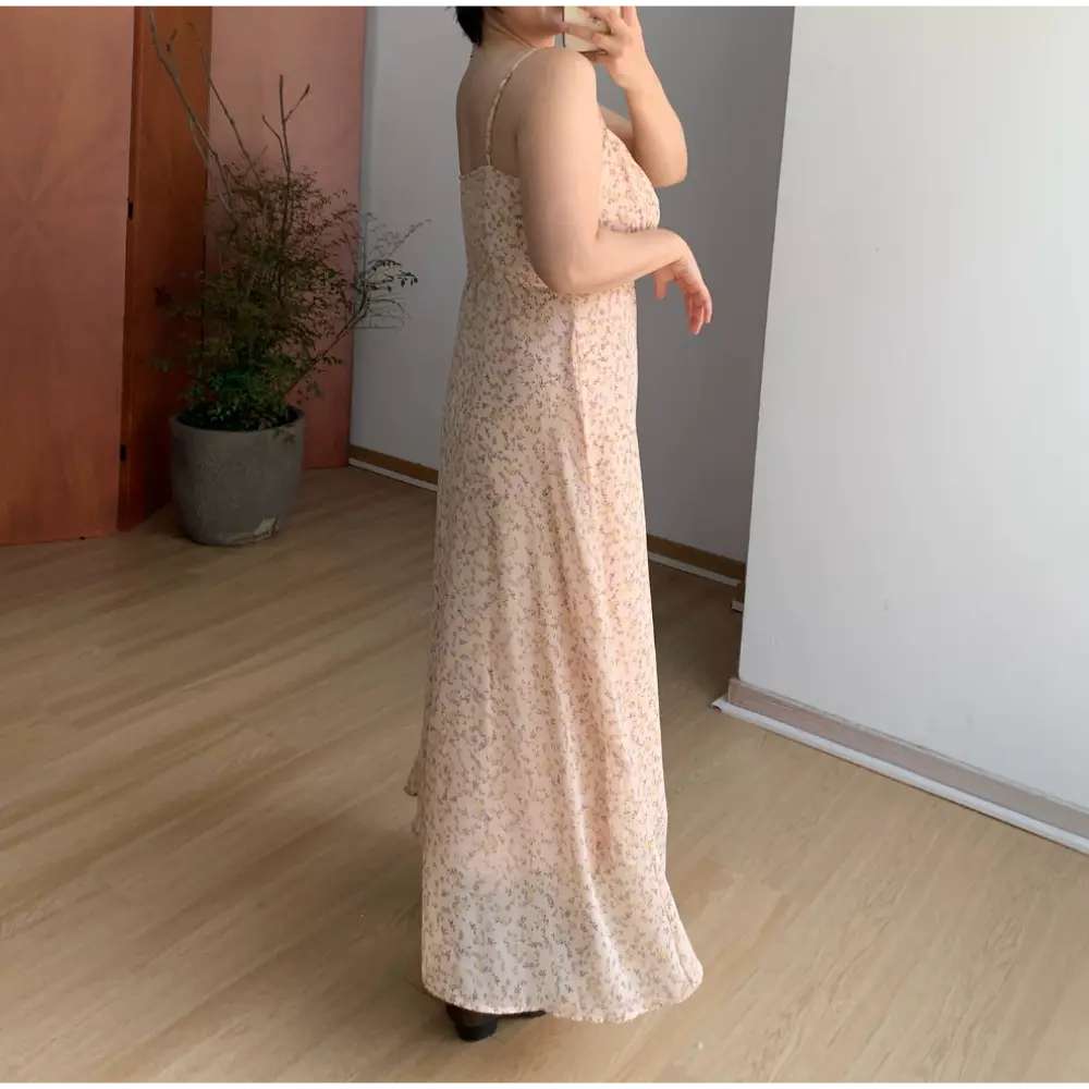 Three-quarter back indoor mirror shot of pale apricot tiny floral maxi slip dress, thin rear adjustable spaghetti straps, full smocked back waist panel, sheer flowy floor-length skirt