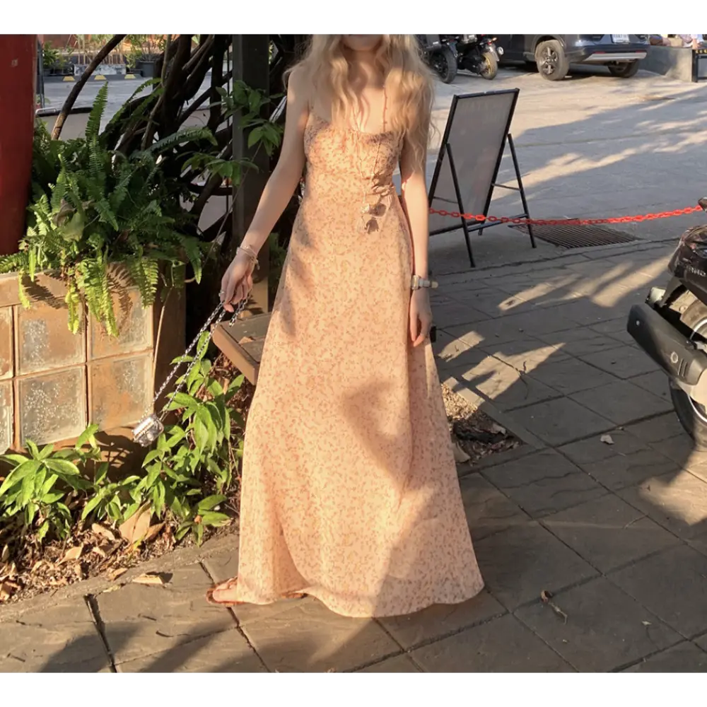 Three-quarter outdoor sidewalk lifestyle shot of pale apricot ditsy floral maxi slip dress, model holding mini silver chain crossbody bag, soft natural sunset light filtering through surrounding green plants