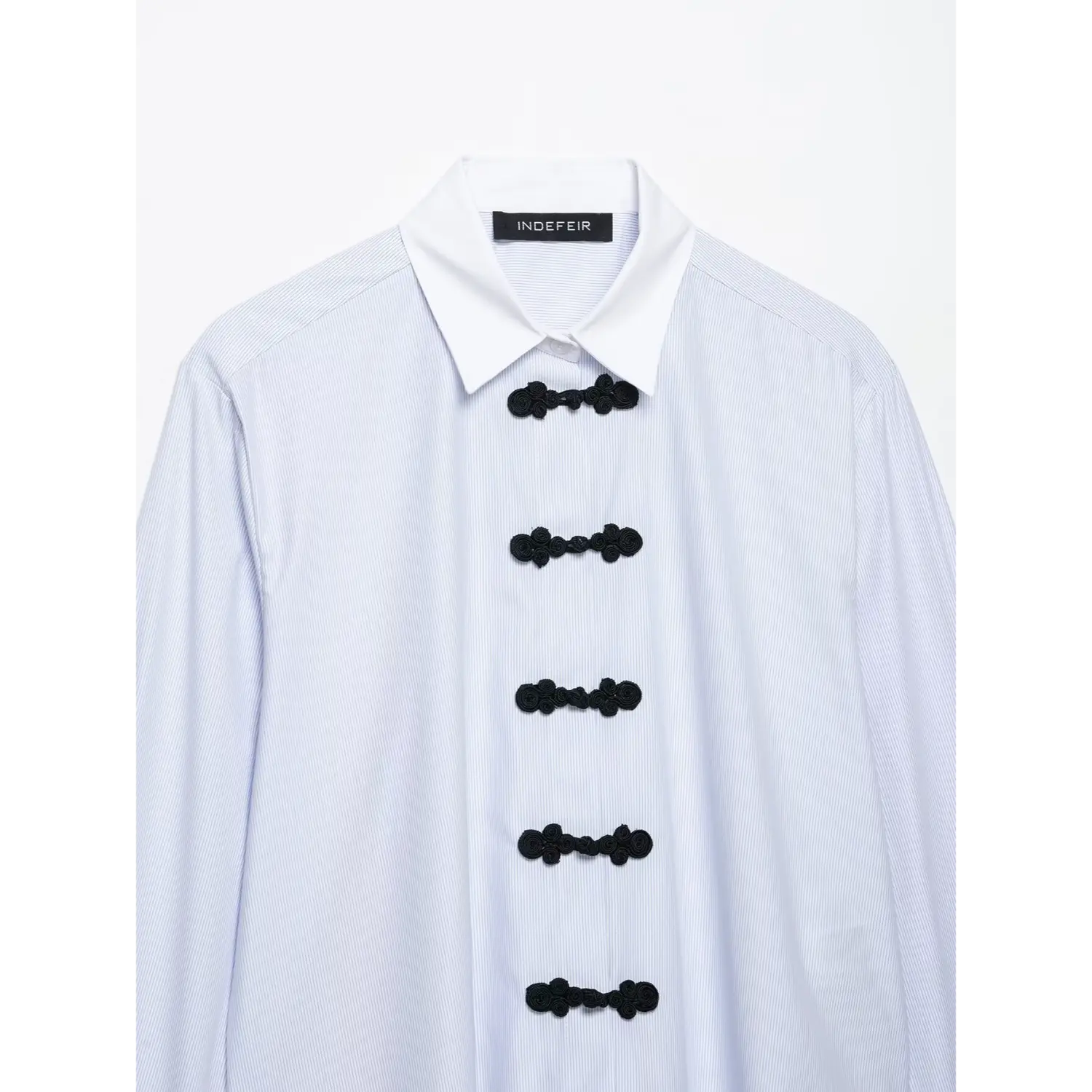 Partial close-up of shirt front, white point collar and vertical row black chinese frog knot buttons on blue white pinstripe woven fabric