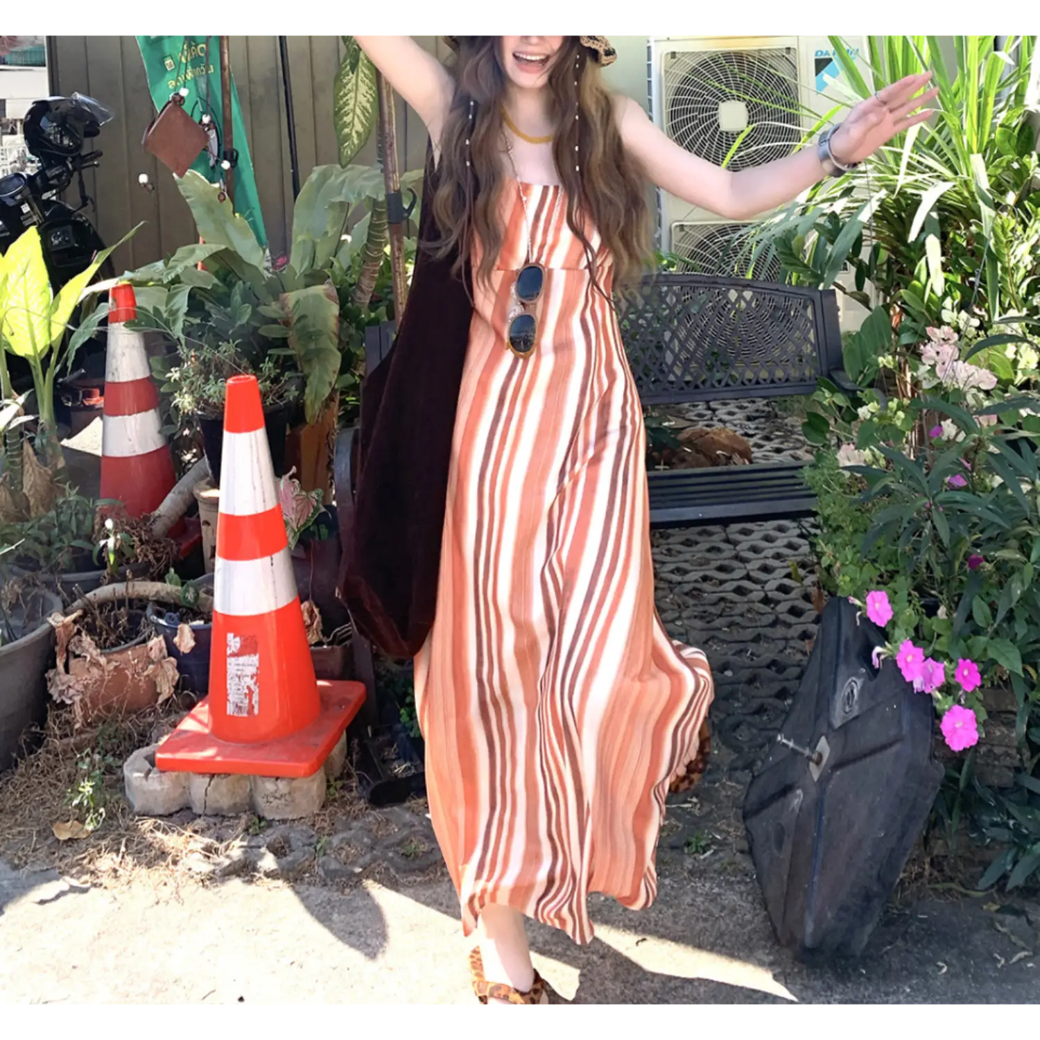 Sunny outdoor courtyard lifestyle shot of warm brown orange striped slip maxi dress, sunglasses hung on bust, model waving arms surrounded by tropical green plants and traffic cones