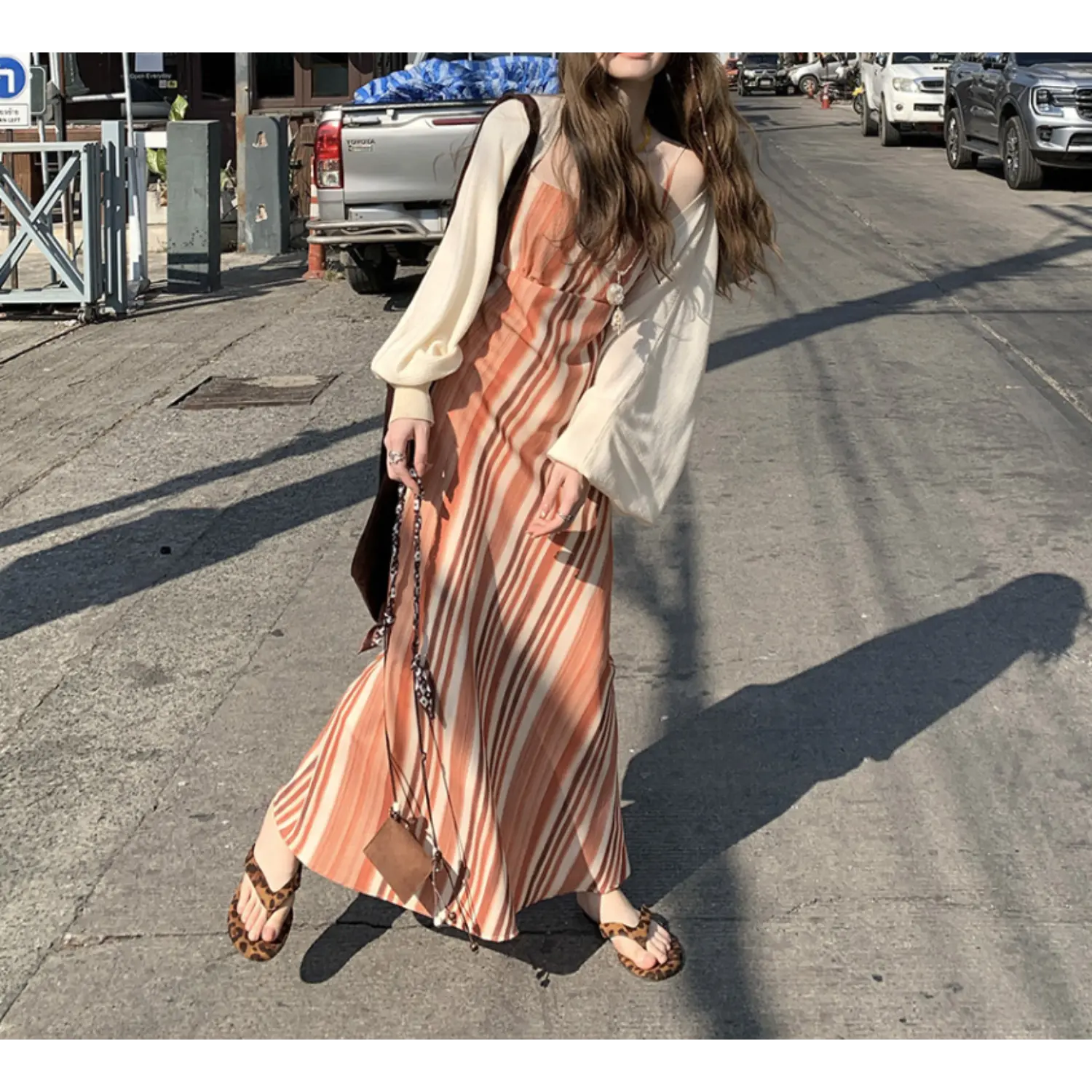 Urban street outdoor lifestyle three-quarter shot of warm tone vertical striped slip maxi dress, cream balloon sleeve layering cardigan, model holding mini leather crossbody accessory