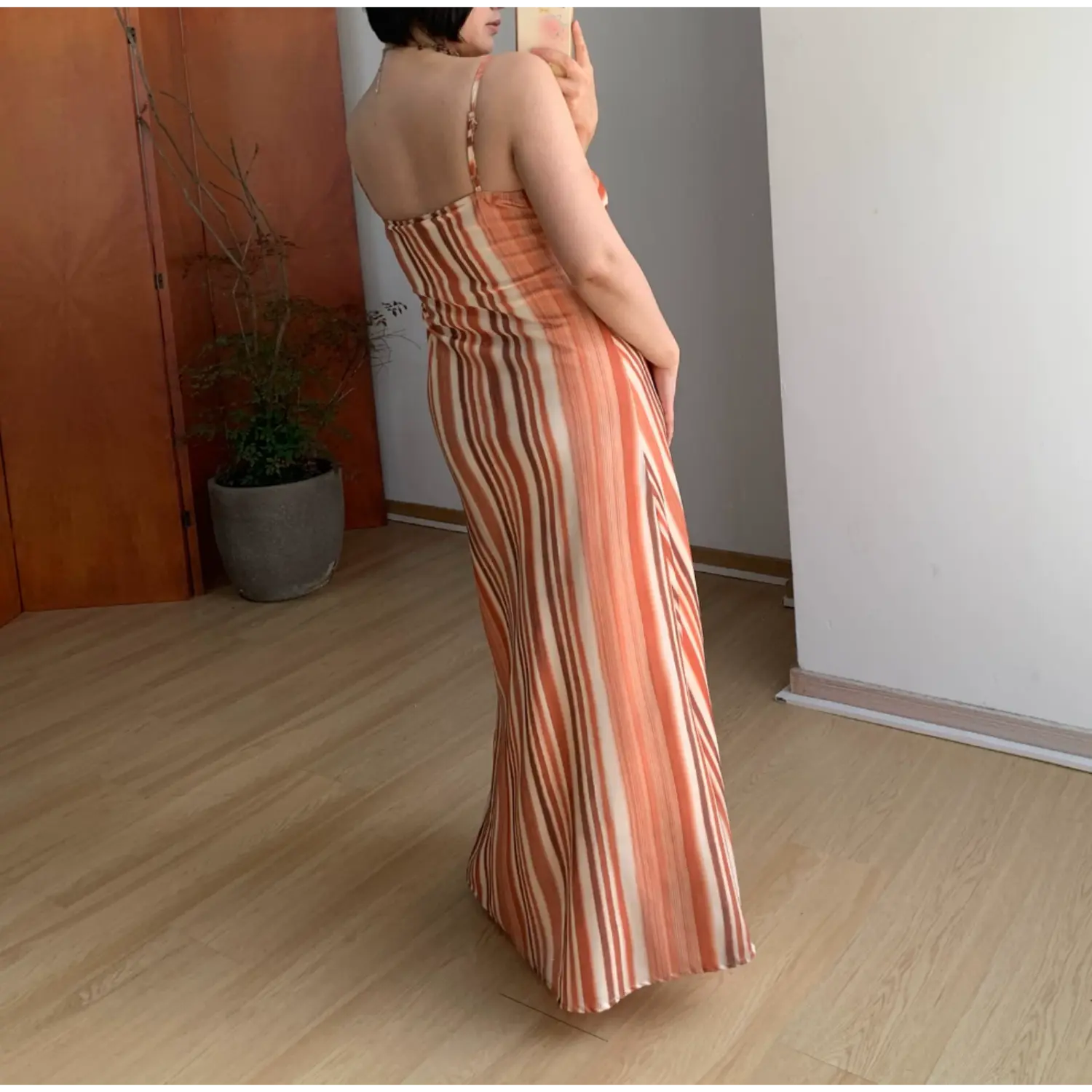 Three-quarter back indoor mirror shot of warm orange brown vertical stripe maxi slip dress, thin adjustable rear spaghetti straps, smooth draped floor-length A-line skirt