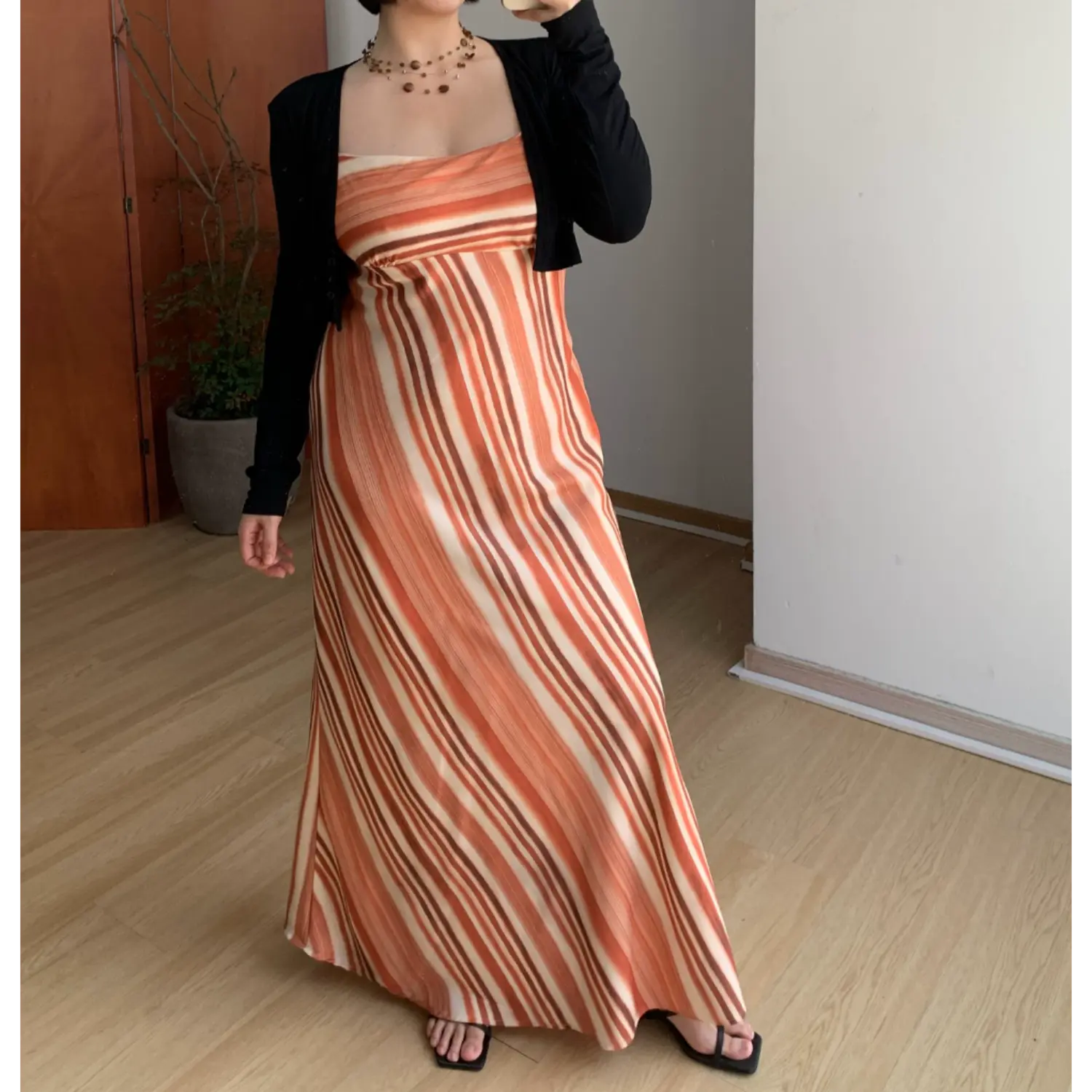 Medium full indoor mirror lifestyle shot of warm tone vertical striped slip maxi dress, layered black fitted cropped knit cardigan, model resting one hand on hip to show slim waist fit