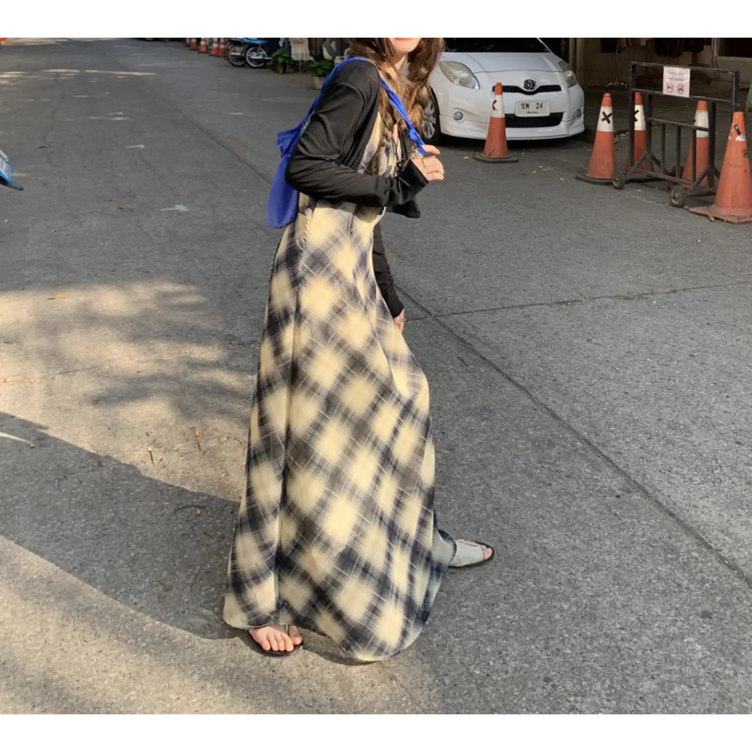 Three-quarter roadside walking shot of sheer chiffon plaid maxi slip dress, cream dark black diagonal check print, slim flared skirt silhouette, thin cami straps, model carrying blue fabric shoulder bag, grey asphalt street with traffic cones & white sedan background