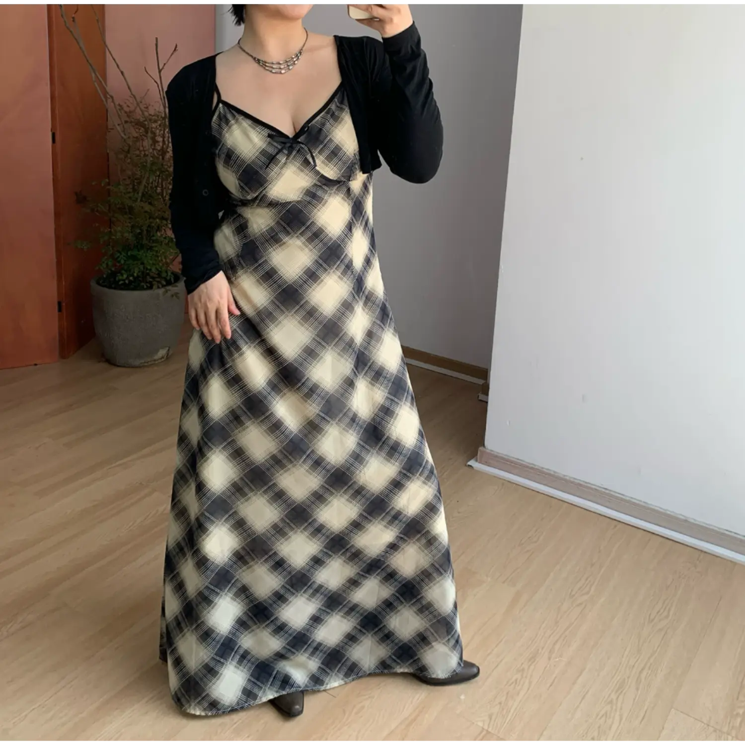 Full indoor mirror shot of bias plaid chiffon maxi slip dress, cream & faded black diagonal check print, thin adjustable spaghetti straps, V-neck with tiny chest bow, black contrast trim, slim flared floor-length skirt, layered with cropped long-sleeve black knit cardigan, styled with brown leather low-heel shoes on light wood flooring