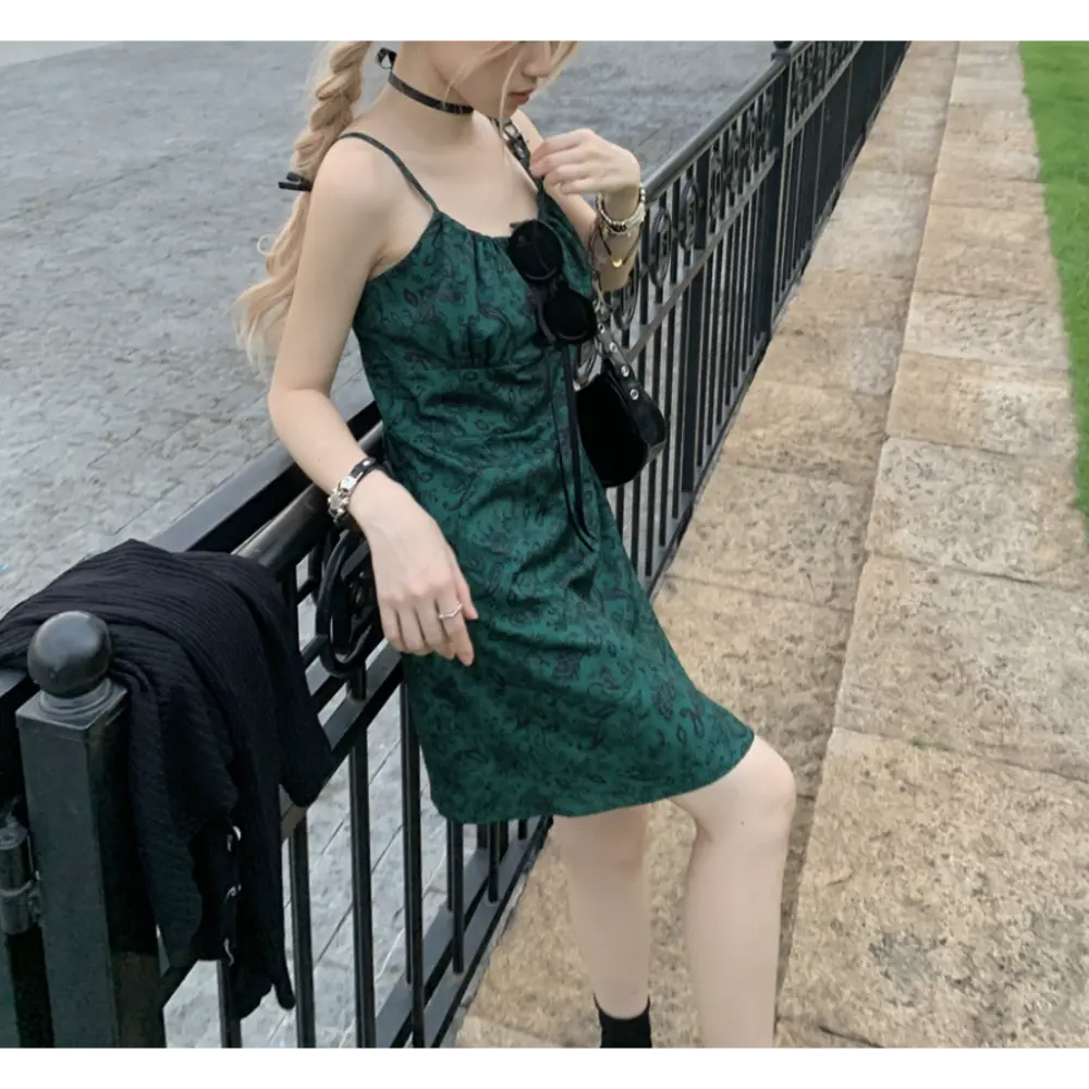 Outdoor sidewalk lifestyle shot of dark green paisley slip mini dress, model leaning on black iron railing, accessorized with black choker, shoulder bag and layered metal bracelets