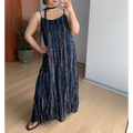 Indoor mirror lifestyle shot of relaxed fit textured slip maxi dress, dark navy vertical bronze-blue faded stripe print, thin cami straps, matching long choker tie, model placing one hand on hip to show loose flowy silhouette, light wood flooring & minimalist white wall background