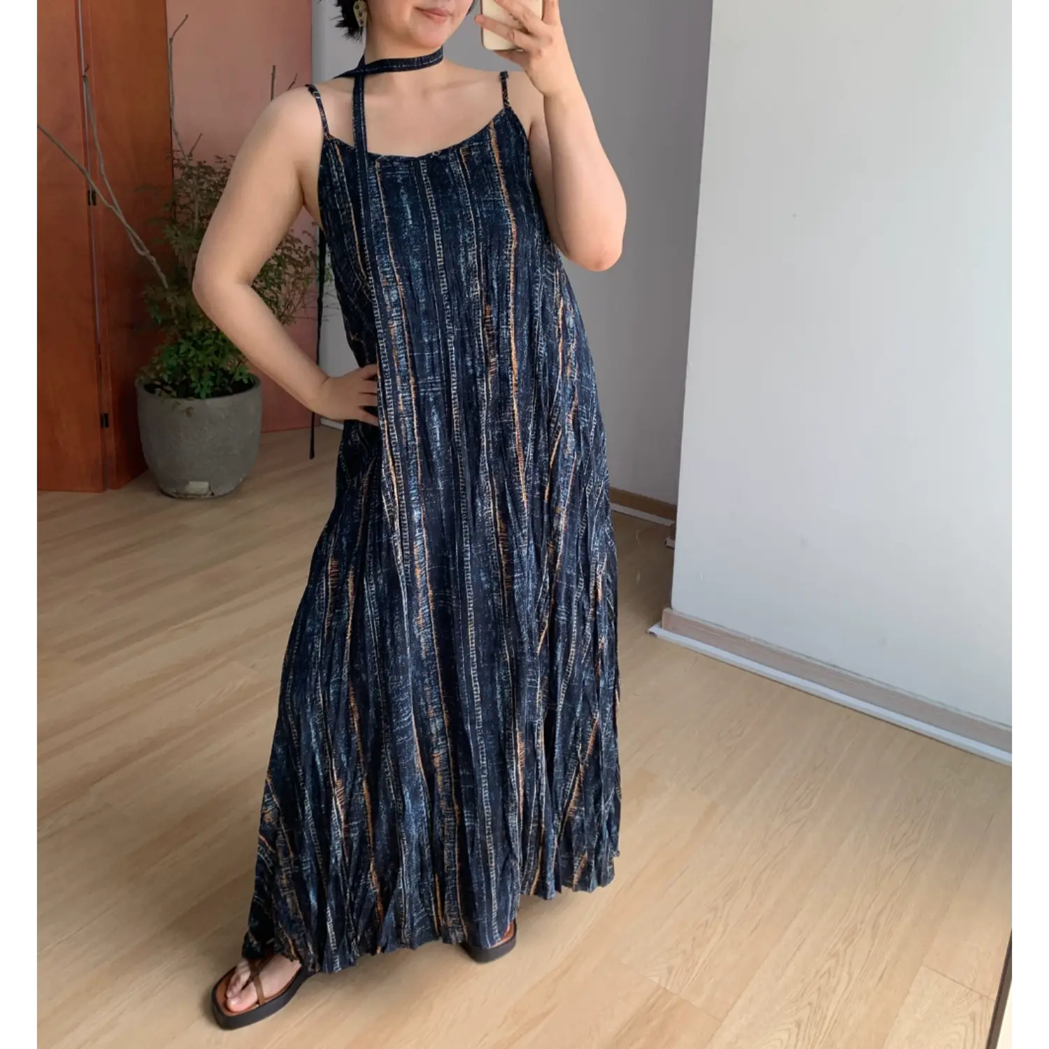 Indoor mirror lifestyle shot of relaxed fit textured slip maxi dress, dark navy vertical bronze-blue faded stripe print, thin cami straps, matching long choker tie, model placing one hand on hip to show loose flowy silhouette, light wood flooring & minimalist white wall background