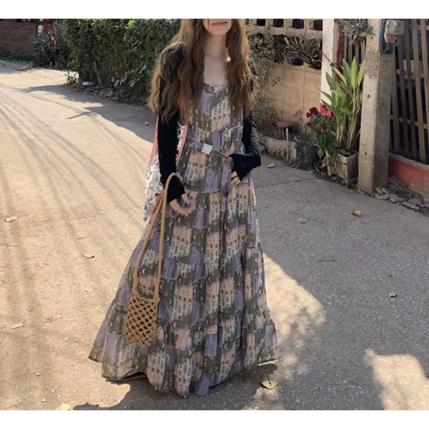 Side outdoor residential street full shot of tier slip maxi dress, muted olive purple collage castle moon print, thin spaghetti straps, loose swing tiered skirt, optional thin waist belt to adjust silhouette, large woven straw shoulder bag, green slide sandals, dry gravel ground & green hedge backdrop