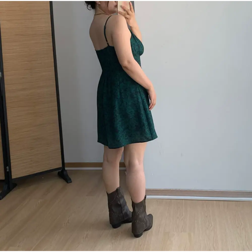 Back view indoor mirror lifestyle shot of dark green paisley cami mini dress, thin rear adjustable straps, fitted waist flared A-line skirt matched with brown western cowboy boots