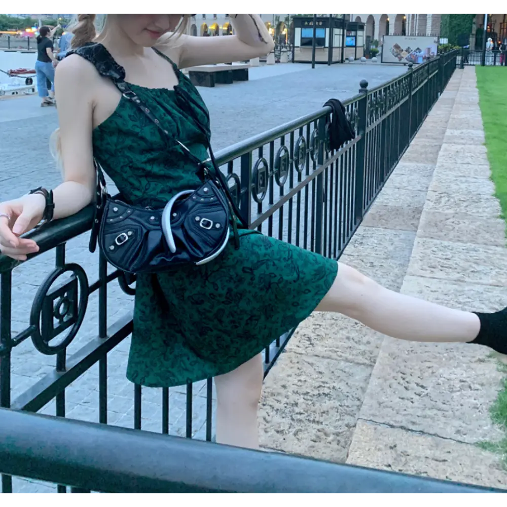 Three-quarter outdoor riverside lifestyle shot of dark green paisley print slip mini dress, model leaning on decorative black iron railing, wearing black ankle socks and vintage moto shoulder bag