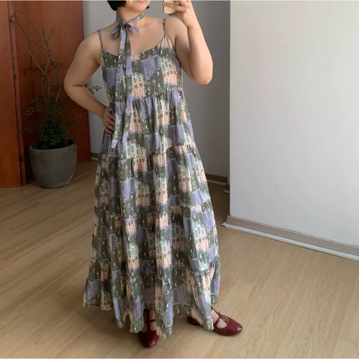 Full indoor mirror shot of loose tiered cami maxi dress, retro collage print with castle, crescent moon & floral motif, muted olive + lavender + cream color palette, thin adjustable spaghetti straps, square scoop neck, matching self-fabric neck tie choker, multi-layer ruffled tier flowy floor skirt, styled with burgundy mary jane flat shoes, minimalist light wood flooring & potted plant decor