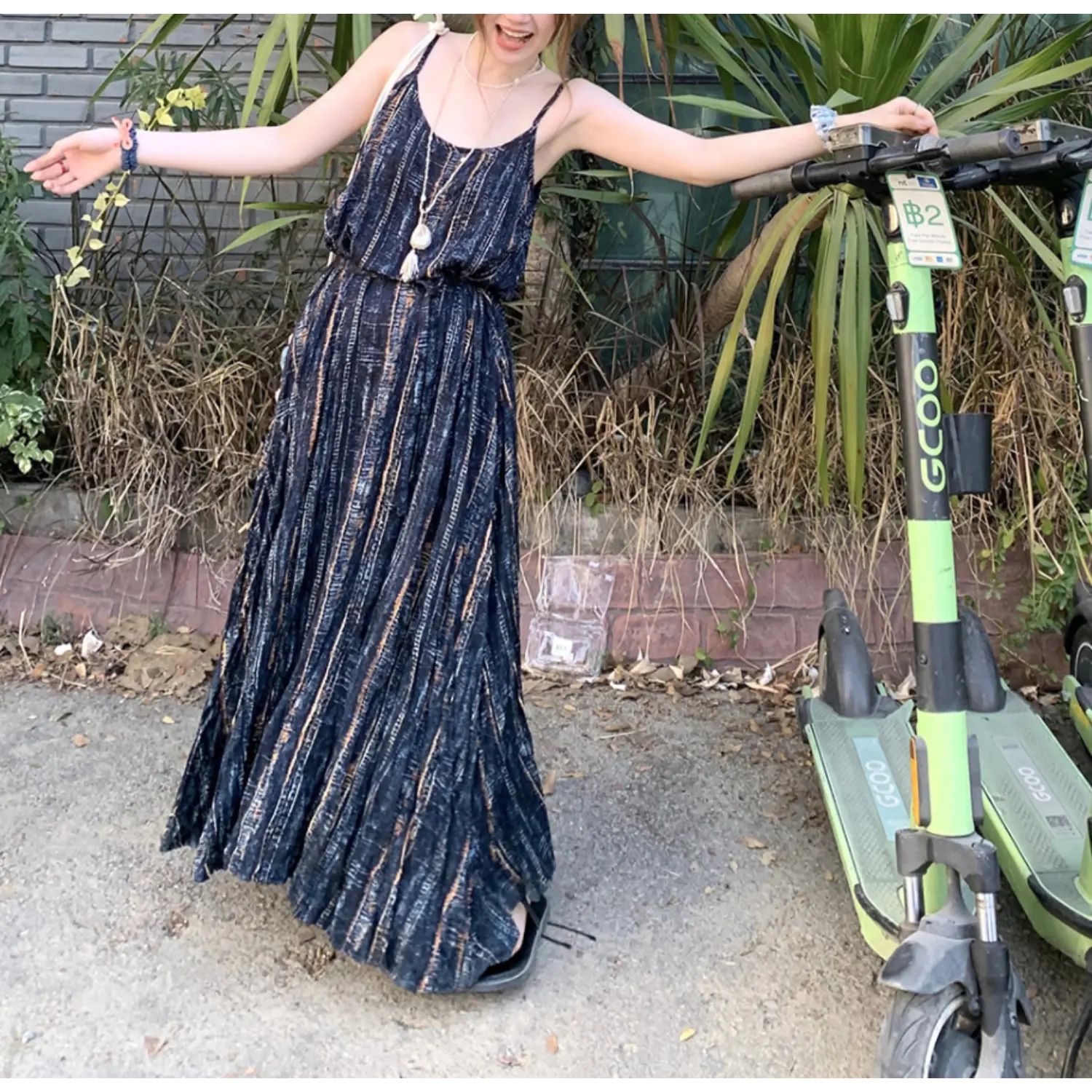 Casual outdoor street lifestyle shot of wrinkle textured cami maxi dress, dark navy faded vertical stripe pattern, relaxed wide swing skirt, thin spaghetti straps, model with arms stretched open beside green shared electric scooter, natural daylight tropical greenery backdrop