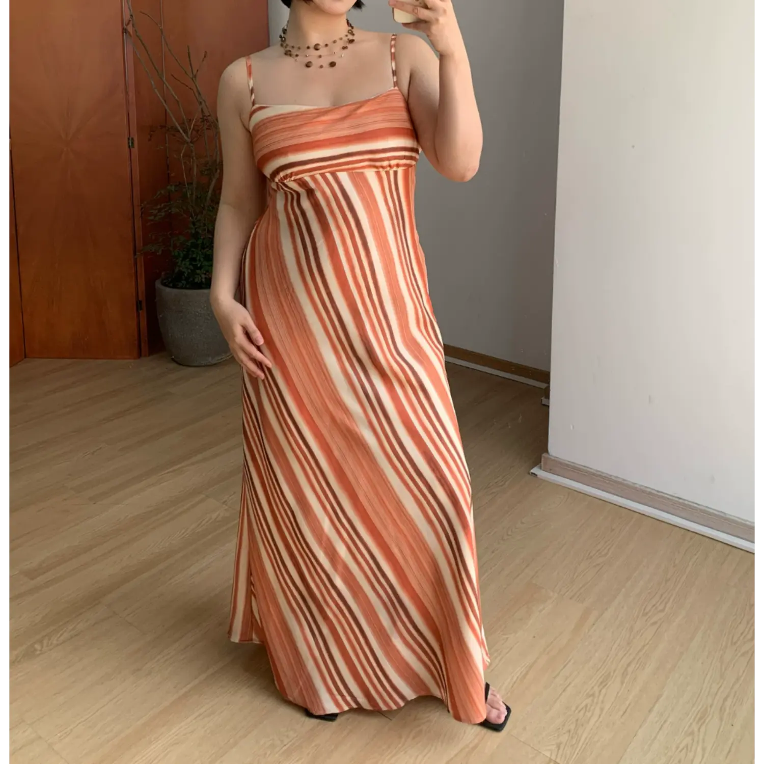Straight front indoor mirror lifestyle photo of terracotta vertical striped slip maxi dress, model gently lifting skirt hem to display lightweight flowy fabric, black square flat sandals