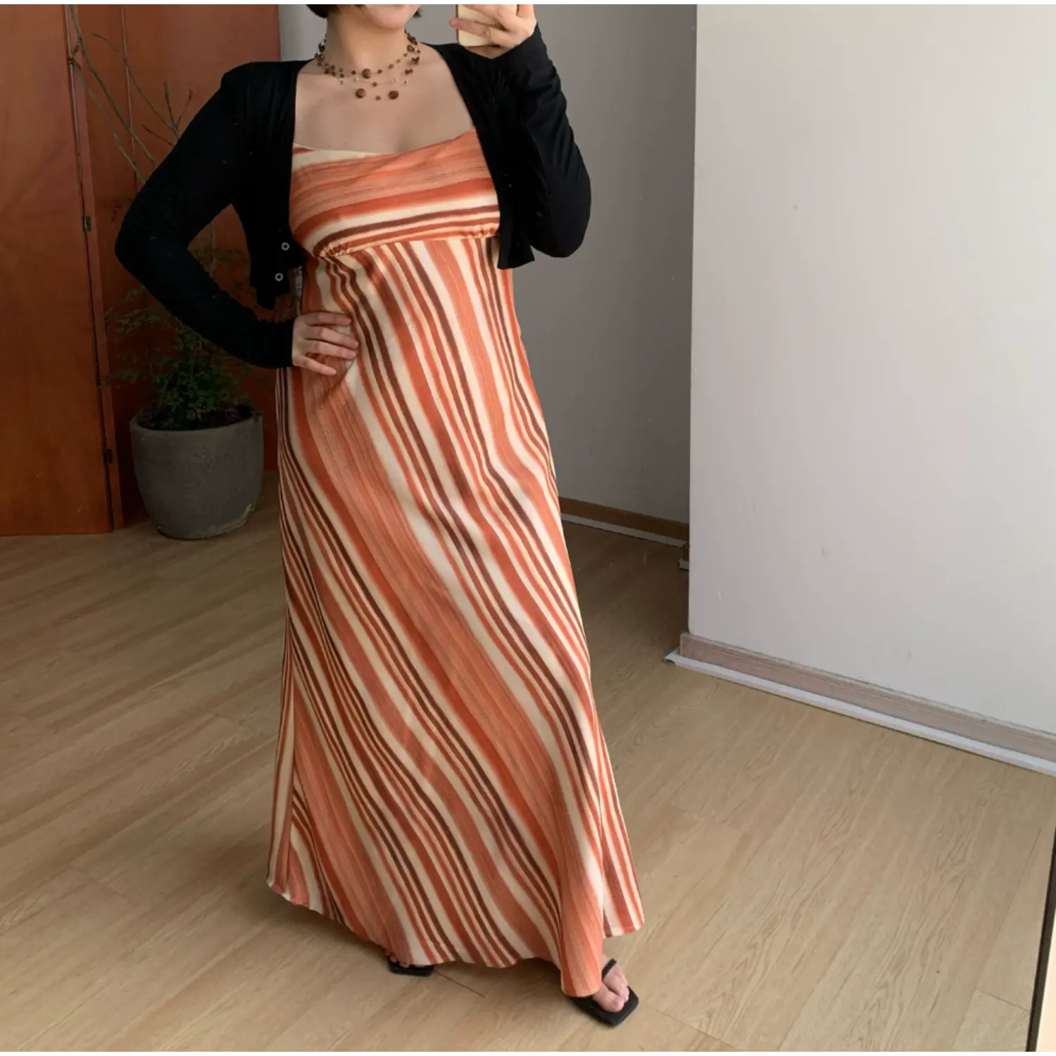 Full body indoor mirror shot of terracotta vertical gradient stripe maxi slip dress, layered slim black long sleeve cropped knit cardigan, matching black square flat slide sandals