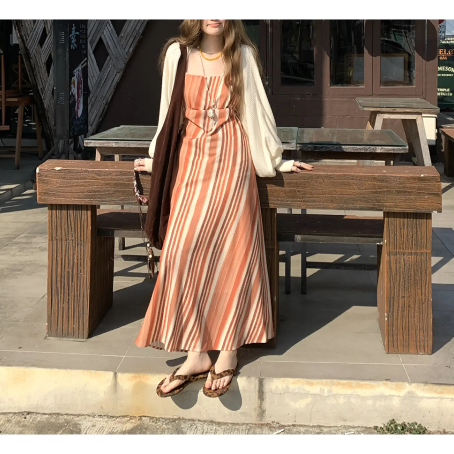 Full body outdoor lifestyle shot of terracotta vertical stripe slip maxi dress, layered cream loose puff sleeve knit cardigan, model leaning on thick solid wooden bench with leopard print flip flops