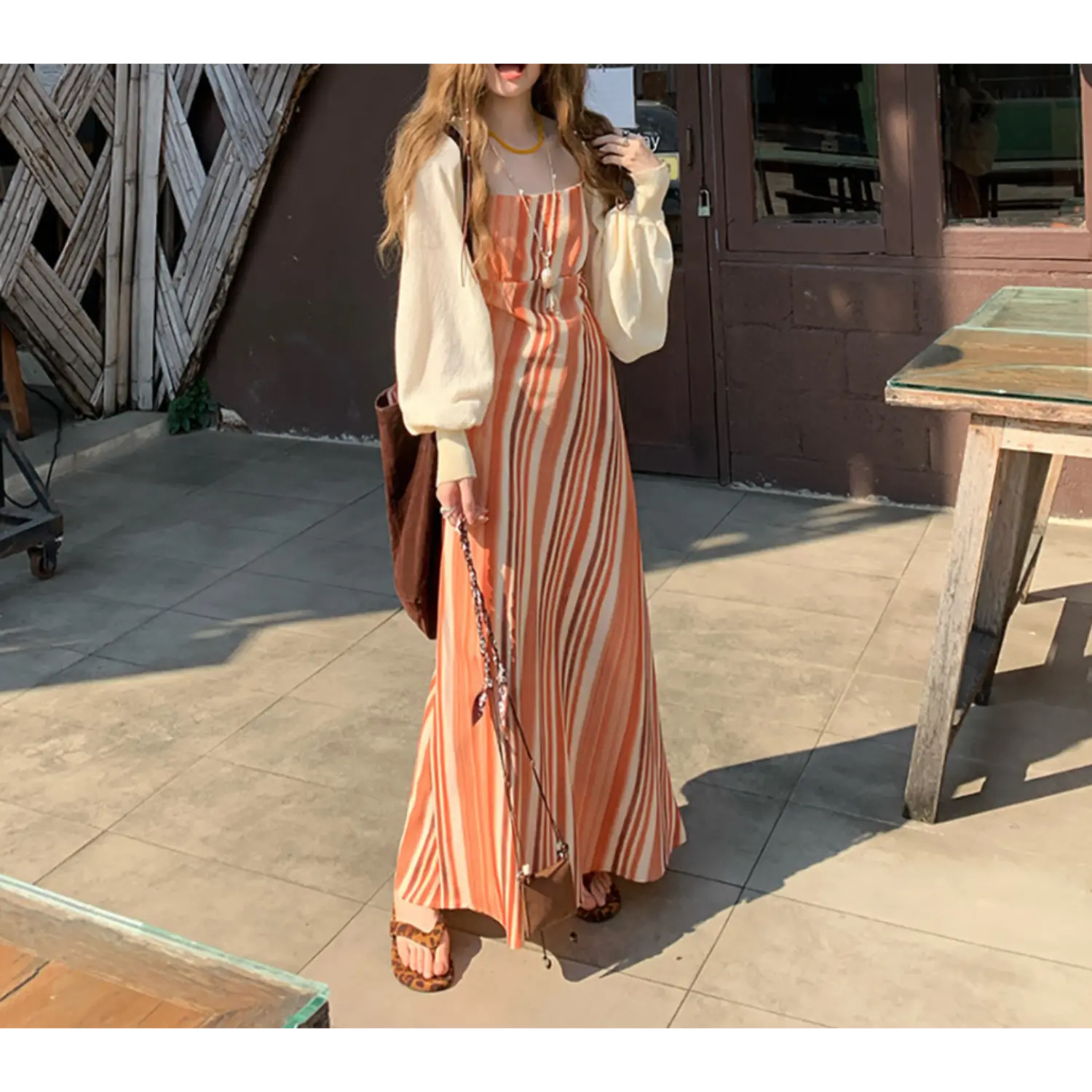 Outdoor storefront lifestyle full shot of terracotta gradient vertical stripe slip maxi dress, thin spaghetti straps, layered oversized cream balloon sleeve knit cardigan, styled with leopard slide sandals and brown canvas tote bag