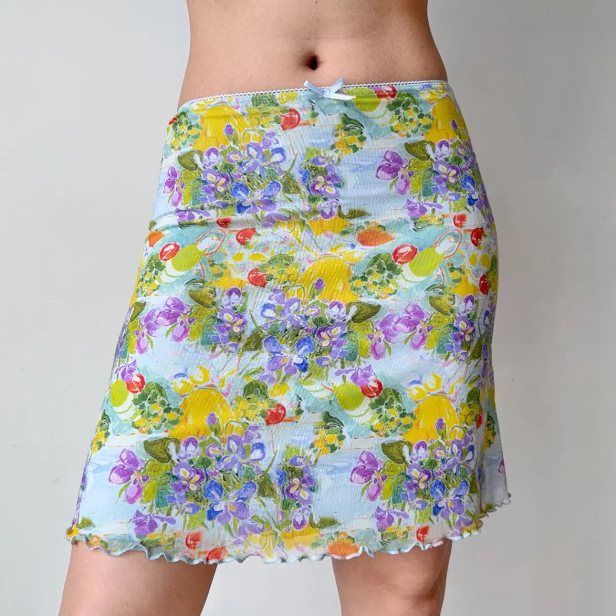 Front view of a light blue mesh mini skirt with a colorful watercolor print of fruits and flowers, showing the delicate waist bow and lettuce-edge hem.
