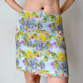 Full view of a light blue mesh mini skirt with a vibrant watercolor print, lettuce-edge hem, and waist bow, worn with a cropped top.