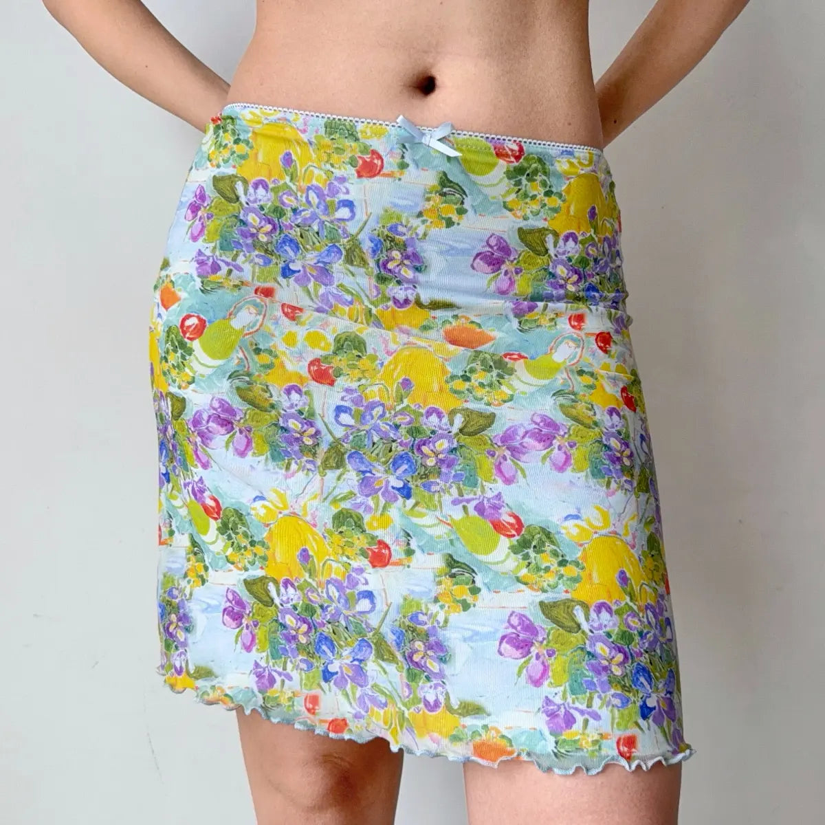 Full view of a light blue mesh mini skirt with a vibrant watercolor print, lettuce-edge hem, and waist bow, worn with a cropped top.